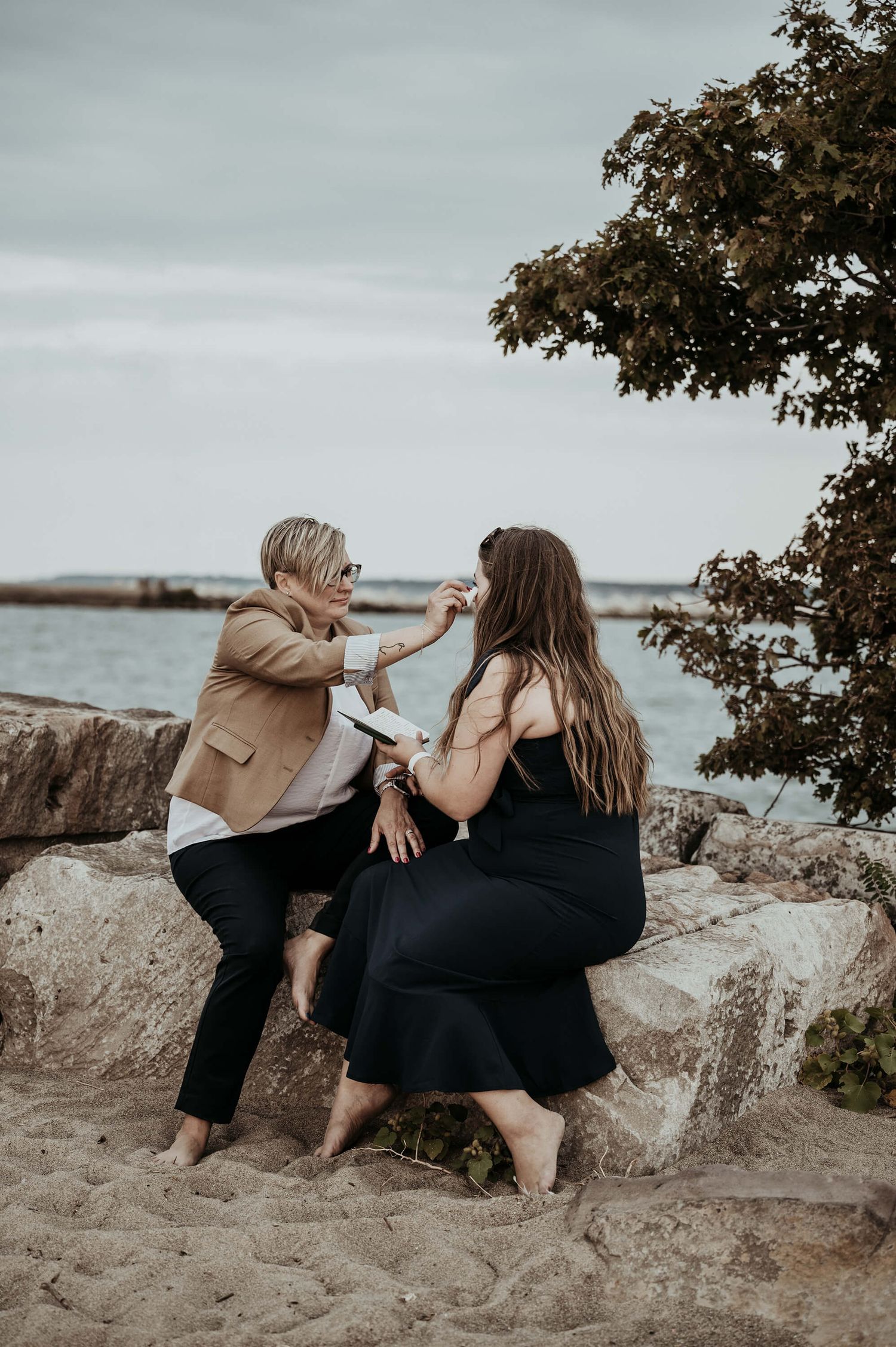 Two people enjoying a candid moment together while sitting on coastal rocks during sunset with ocean views and tree branches above.