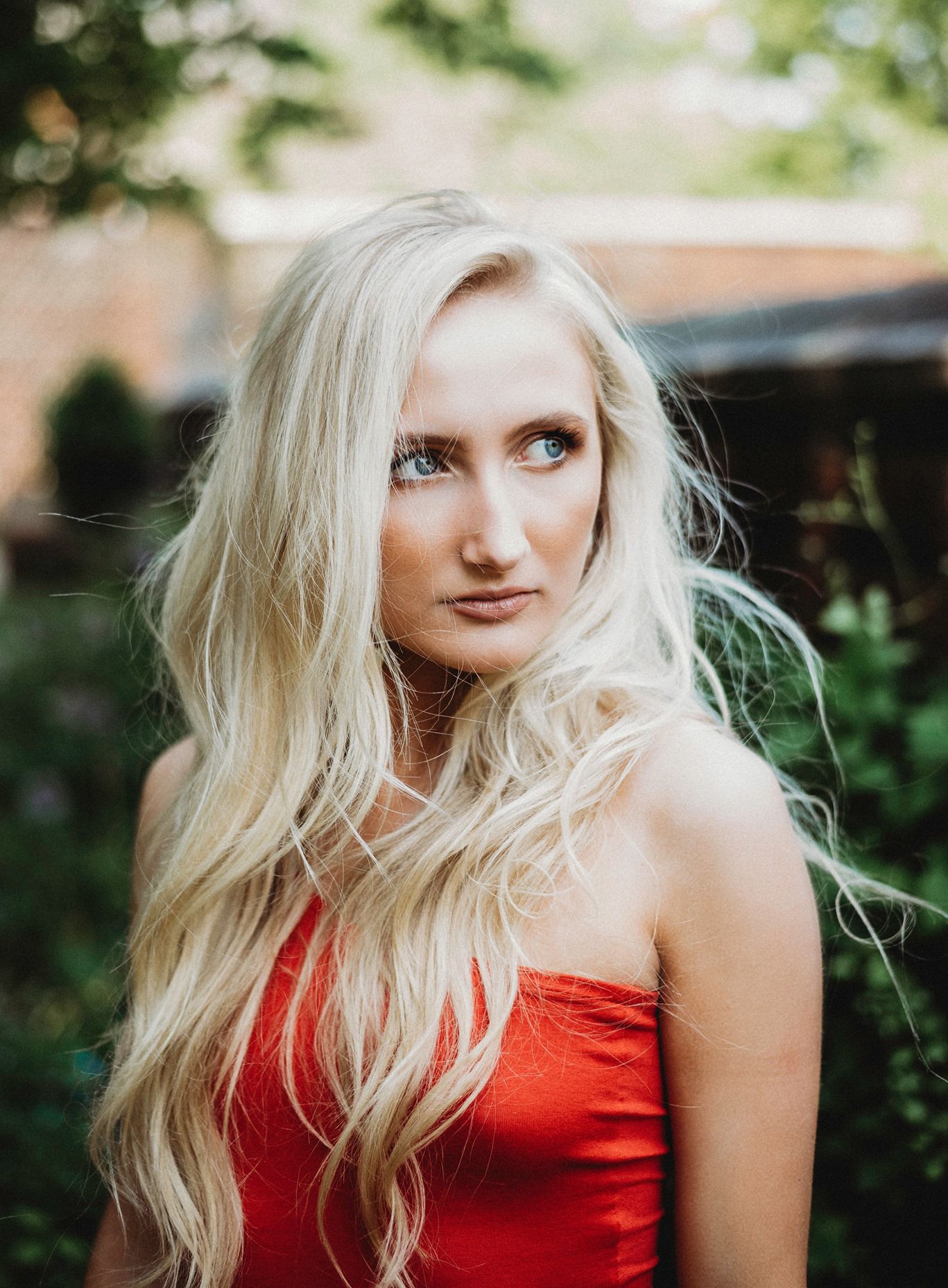 A portrait of someone with long platinum blonde hair wearing a red strapless dress against a soft blurred outdoor background.