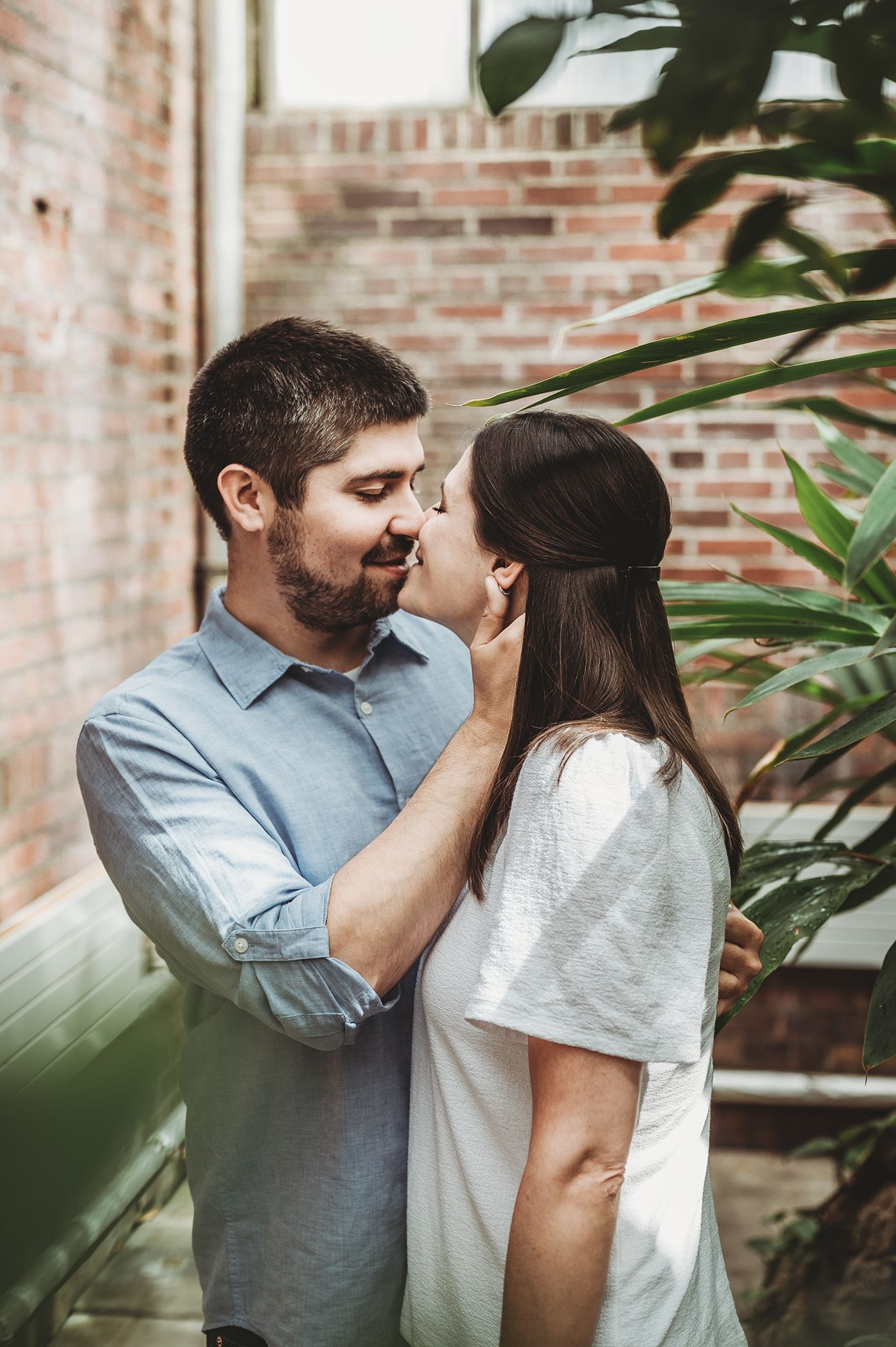 A romantic couple shares a tender moment in front of a brick wall surrounded by green plants.