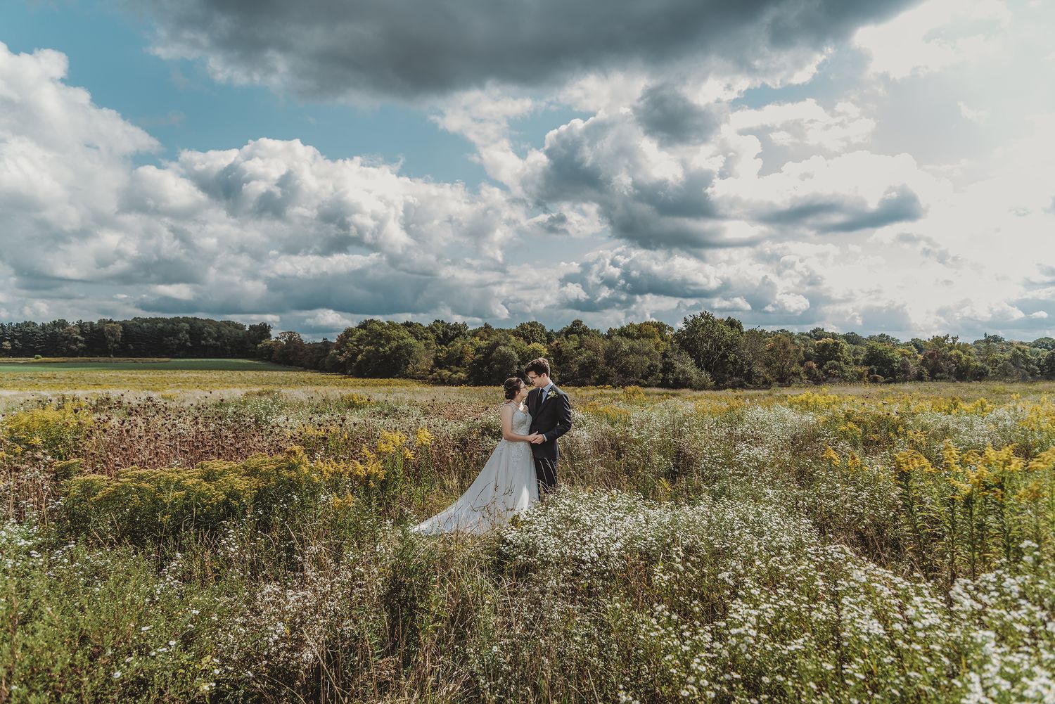A couple stands together in a sunny meadow filled with wildflowers under dramatic cloudy skies.