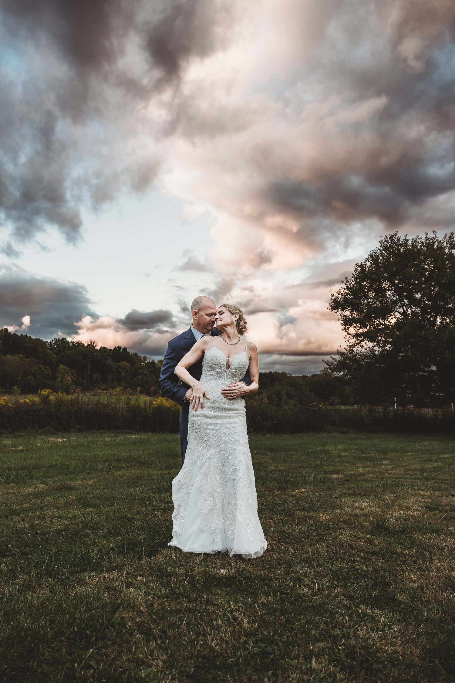 A romantic moment captured outdoors at sunset with dramatic stormy clouds and a lace wedding dress in a grassy field.