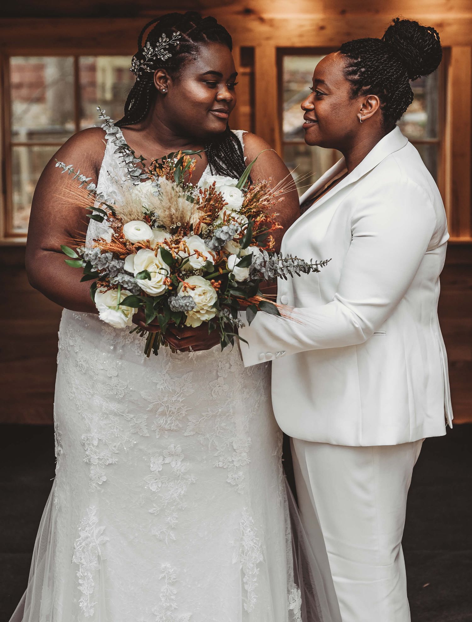 A romantic wedding portrait showing a couple in white embracing in a rustic indoor setting with a beautiful floral bouquet.