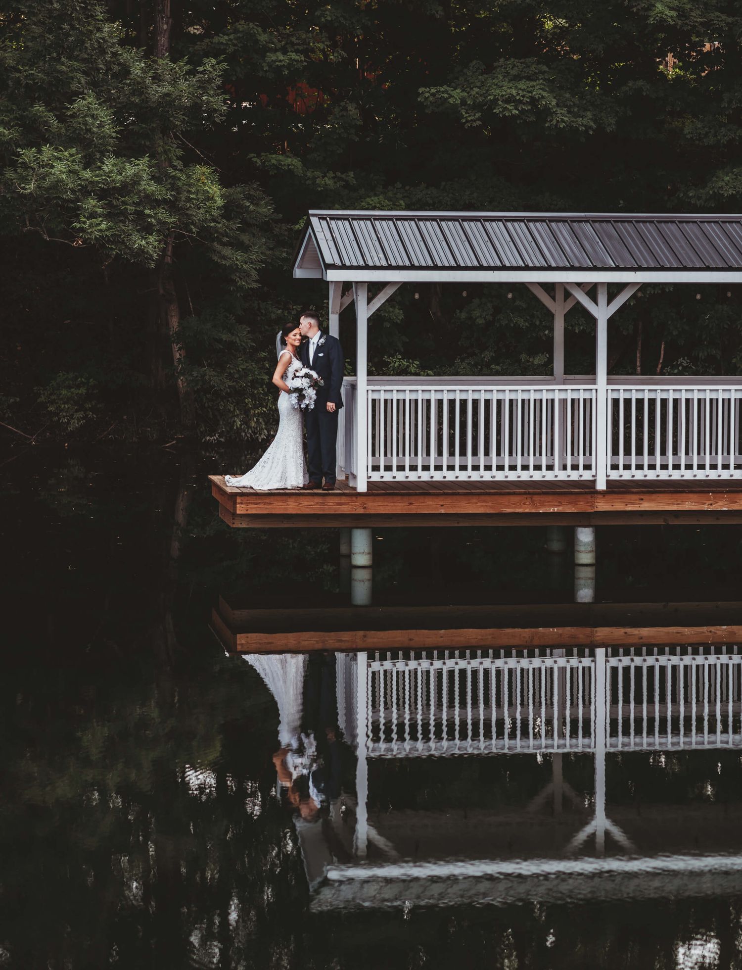 A couple sharing a romantic moment on a covered wooden gazebo dock with a perfect reflection in the still water below.