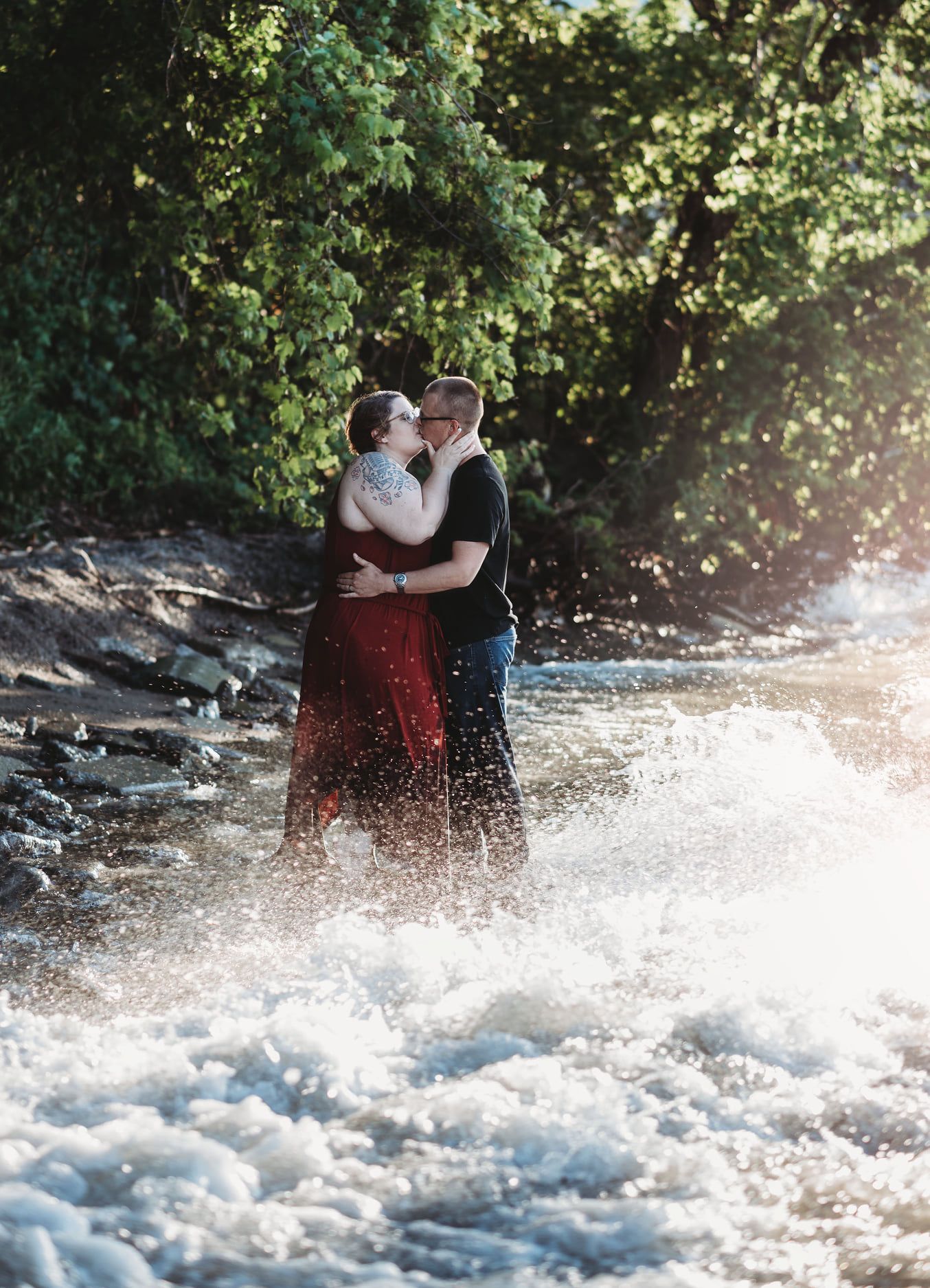 Two people embrace while standing in rushing river water with sunlit trees in the background.