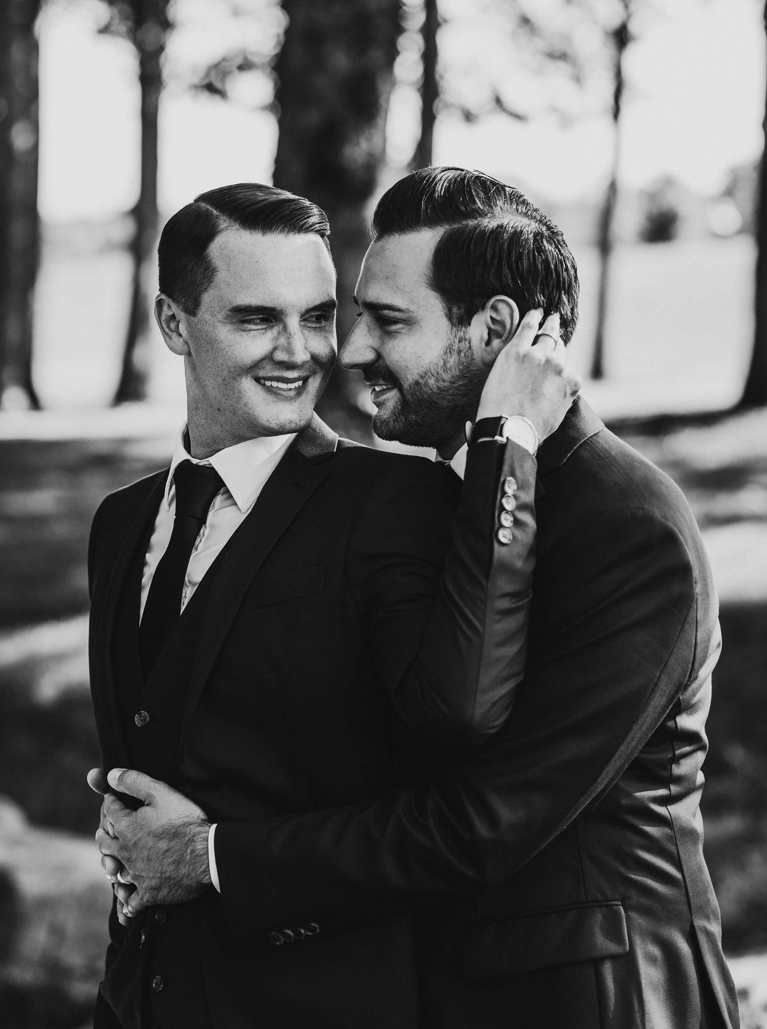 A black and white photograph of a couple in suits sharing an intimate moment and laughing together outdoors.