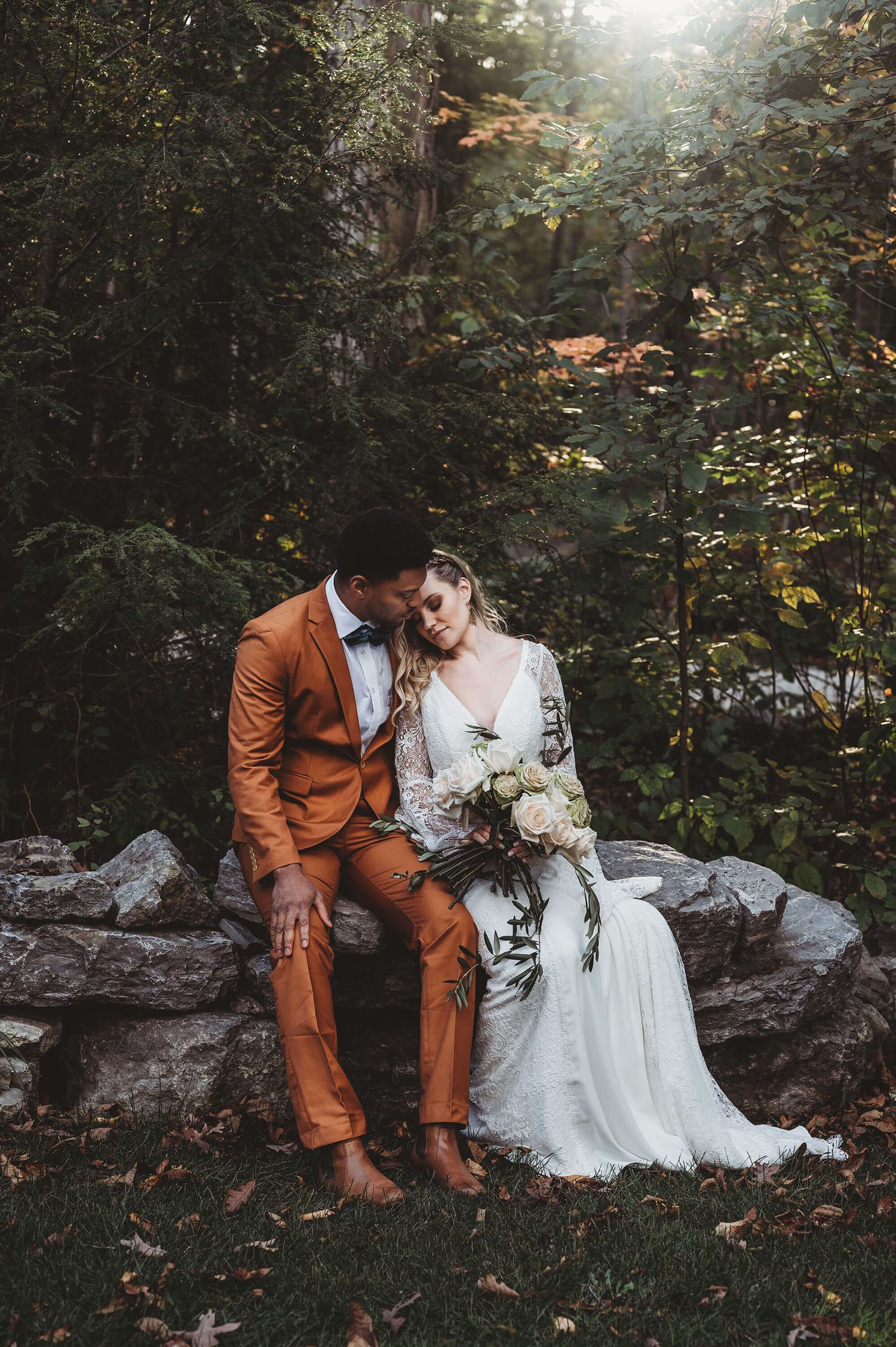 A couple sits together on rocks in a forest setting wearing an orange suit and white wedding dress.