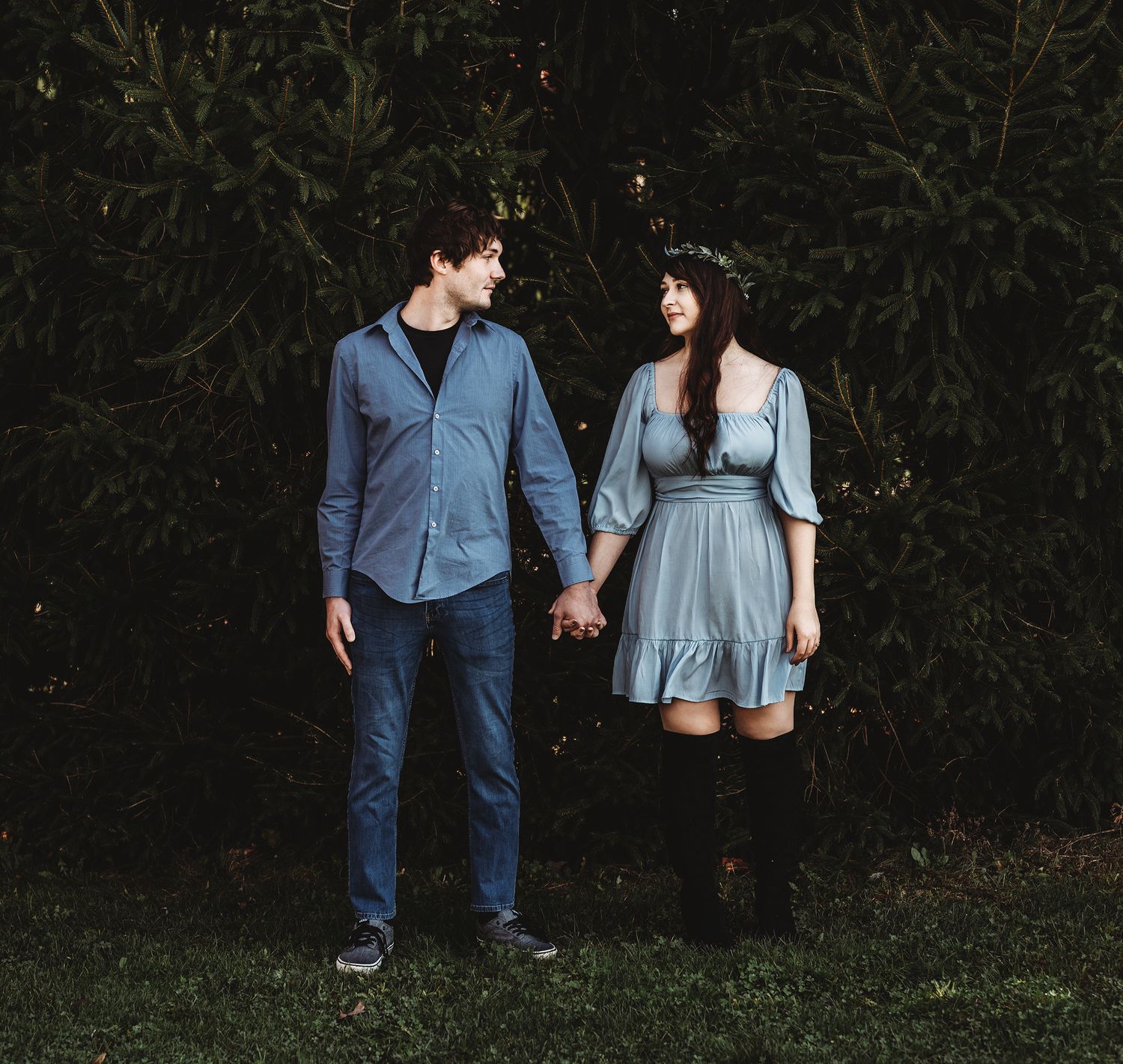 A couple in casual blue outfits holds hands while standing against dark foliage in a romantic outdoor portrait.