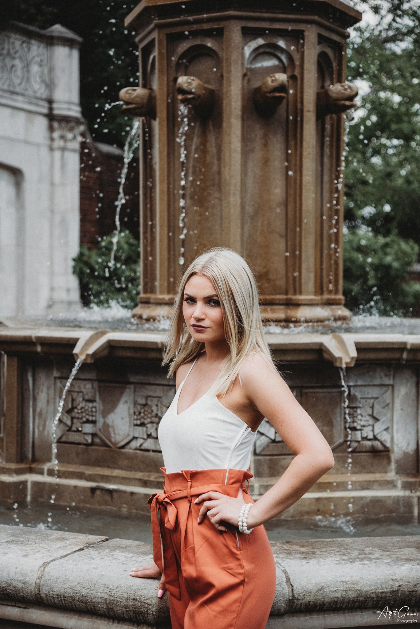 Woman in white and orange dress poses elegantly by ornate stone fountain during outdoor photo session.
