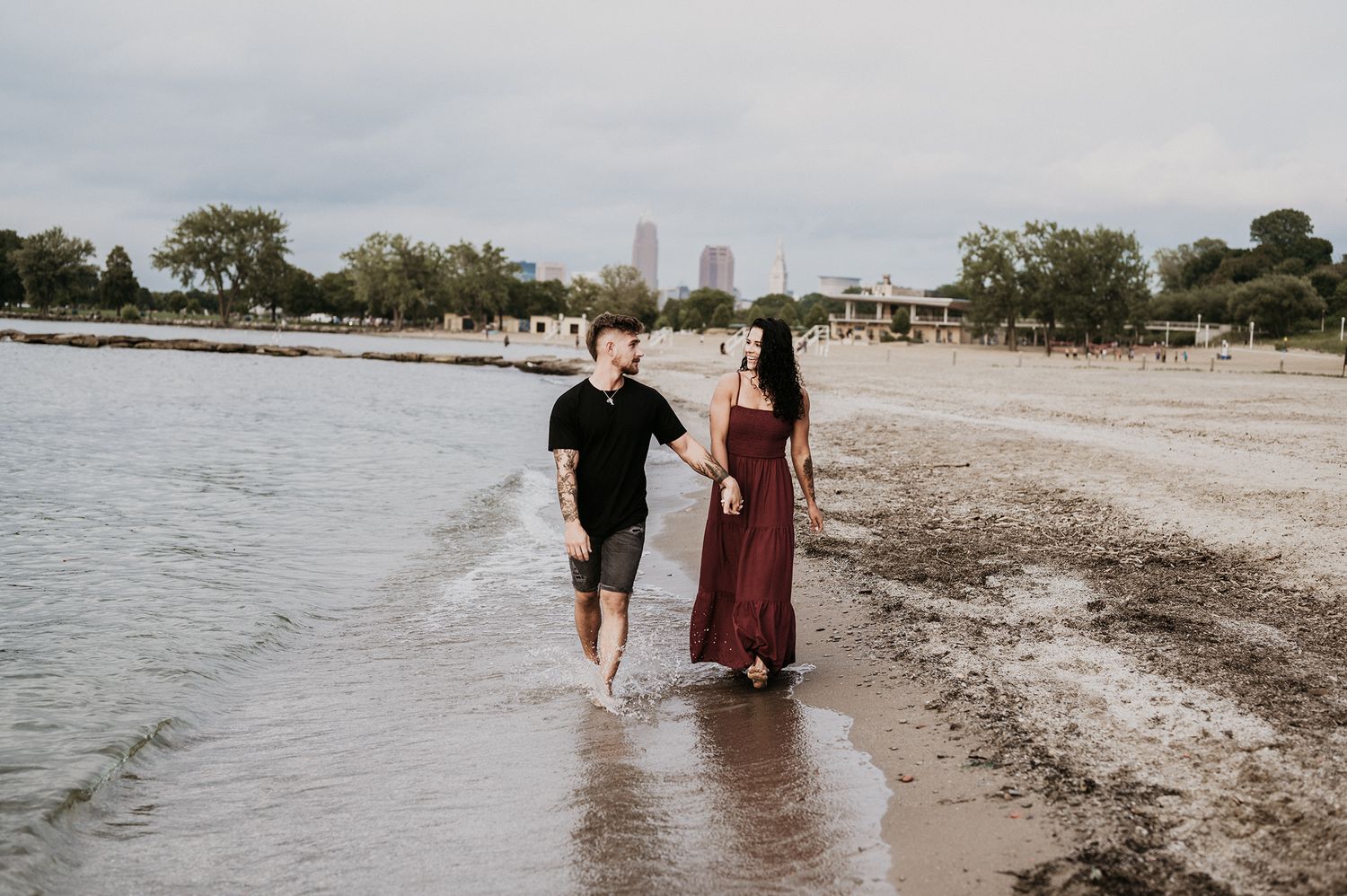 Couple walking barefoot along beach shoreline with city skyline in background.