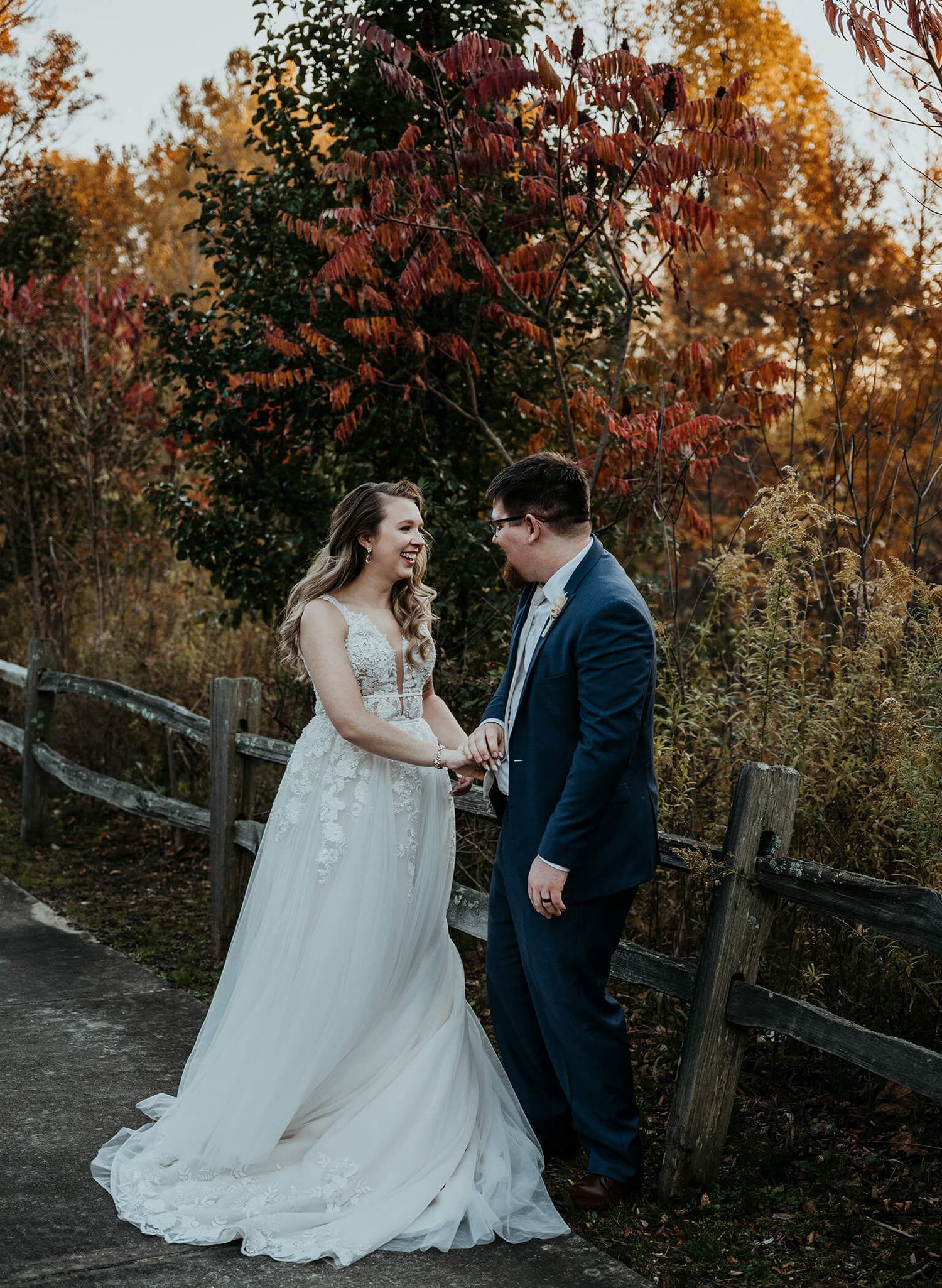 A couple in wedding attire stand together on a path surrounded by vibrant autumn foliage and rustic fencing.