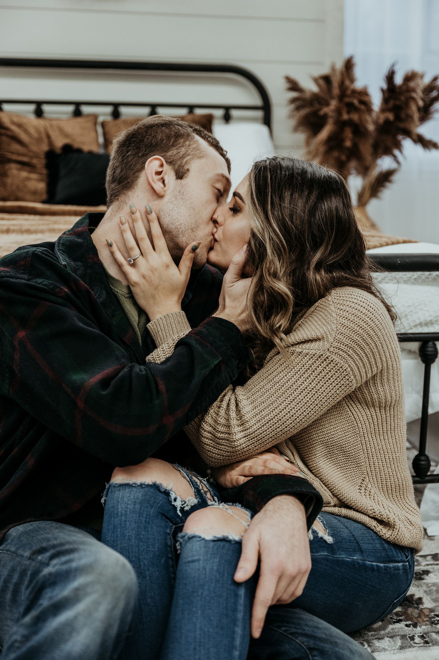 Couple in casual sweaters share an intimate kiss while sitting on floor by metal bed frame with dried pampas grass decor.