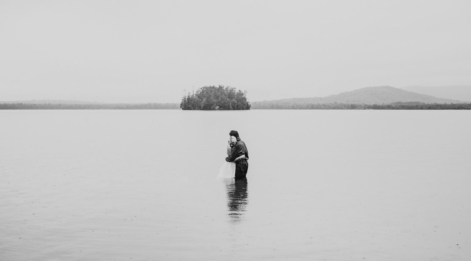 Solitary figure fishing in calm water with a small island and distant mountains visible on the horizon.