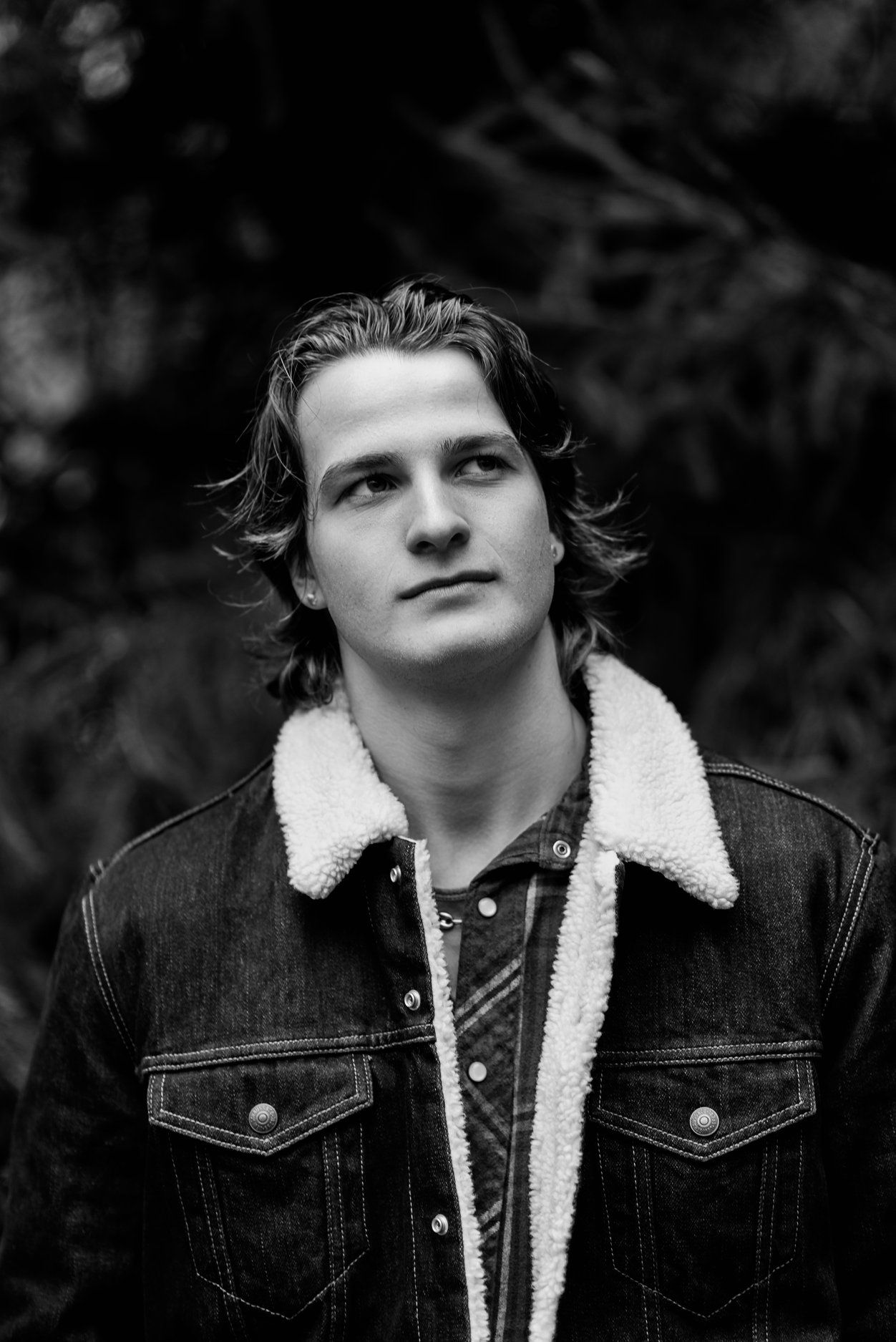 Black and white portrait of someone wearing a denim jacket with sherpa lining against a dark natural background.