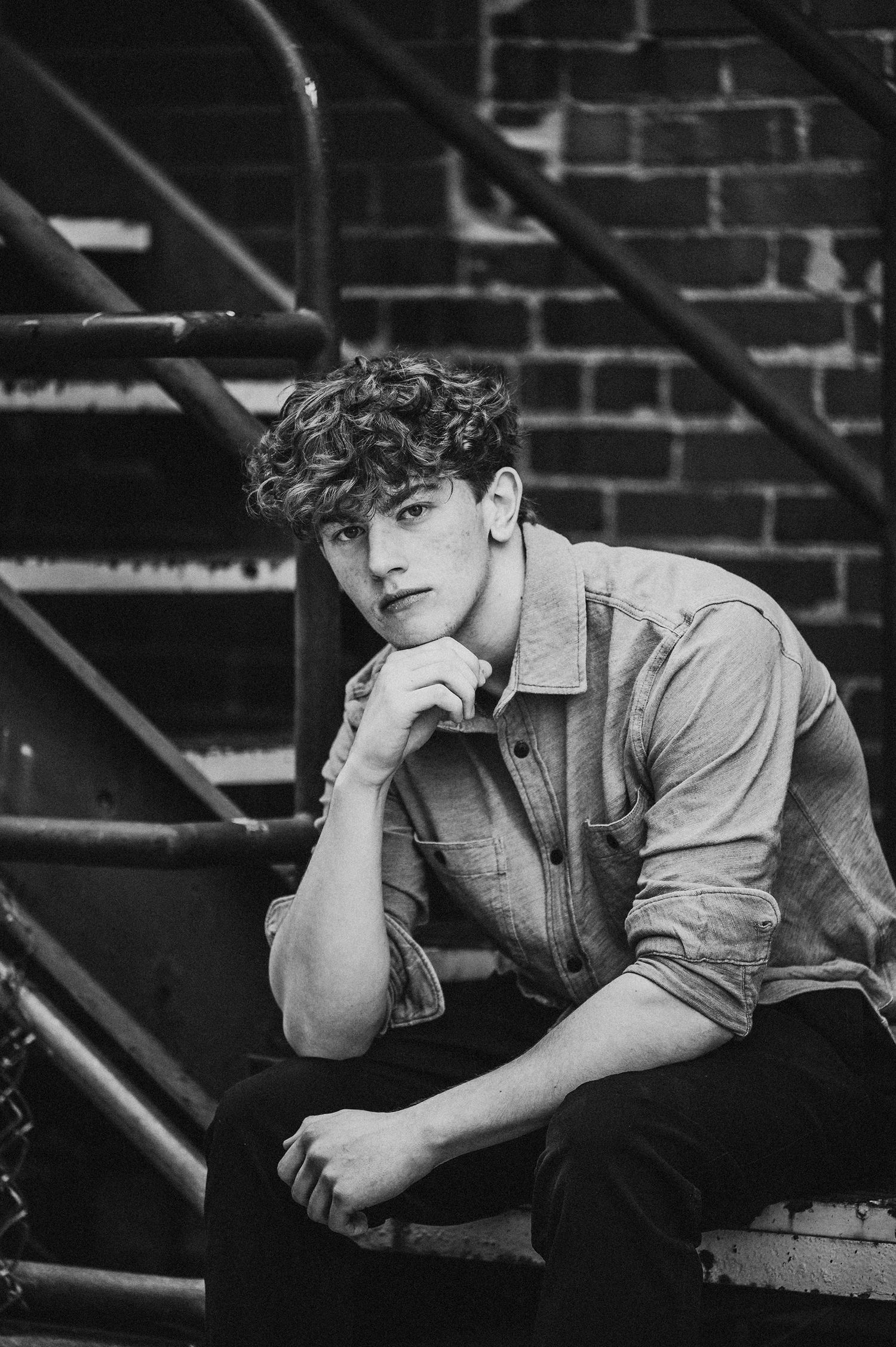 Black and white portrait of someone in casual denim sitting on outdoor stairs against a brick wall background.