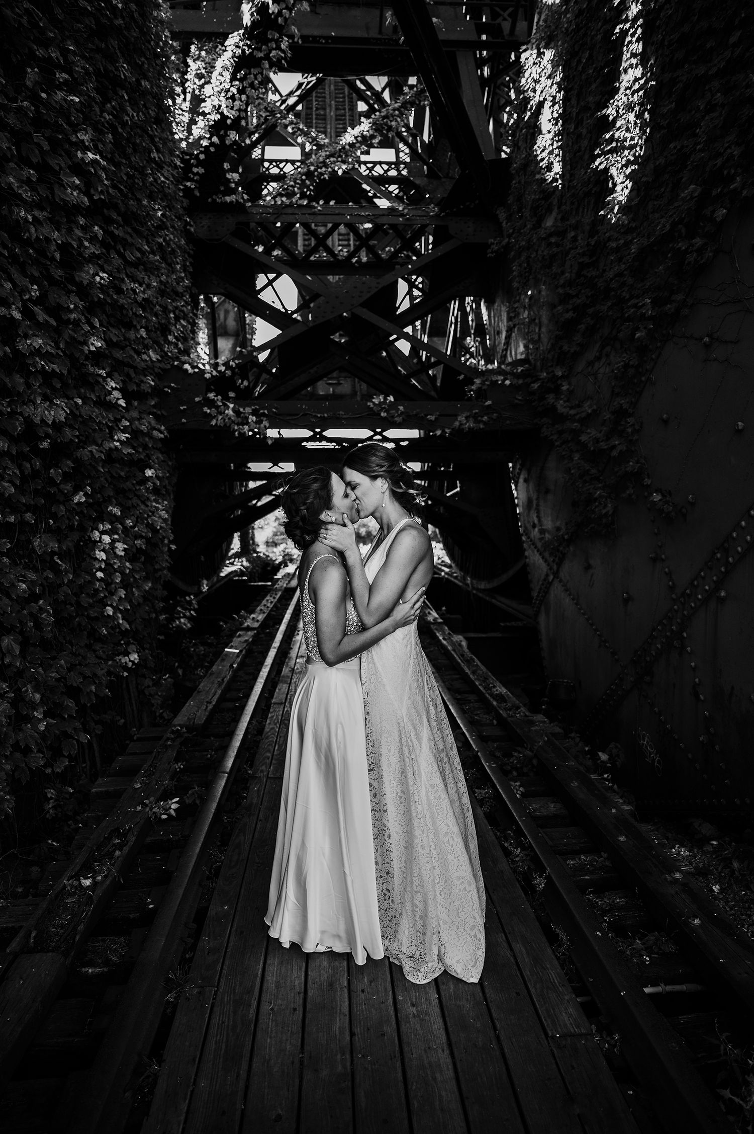 Two people in white wedding attire embrace intimately on a wooden walkway framed by ivy-covered walls and industrial beams.