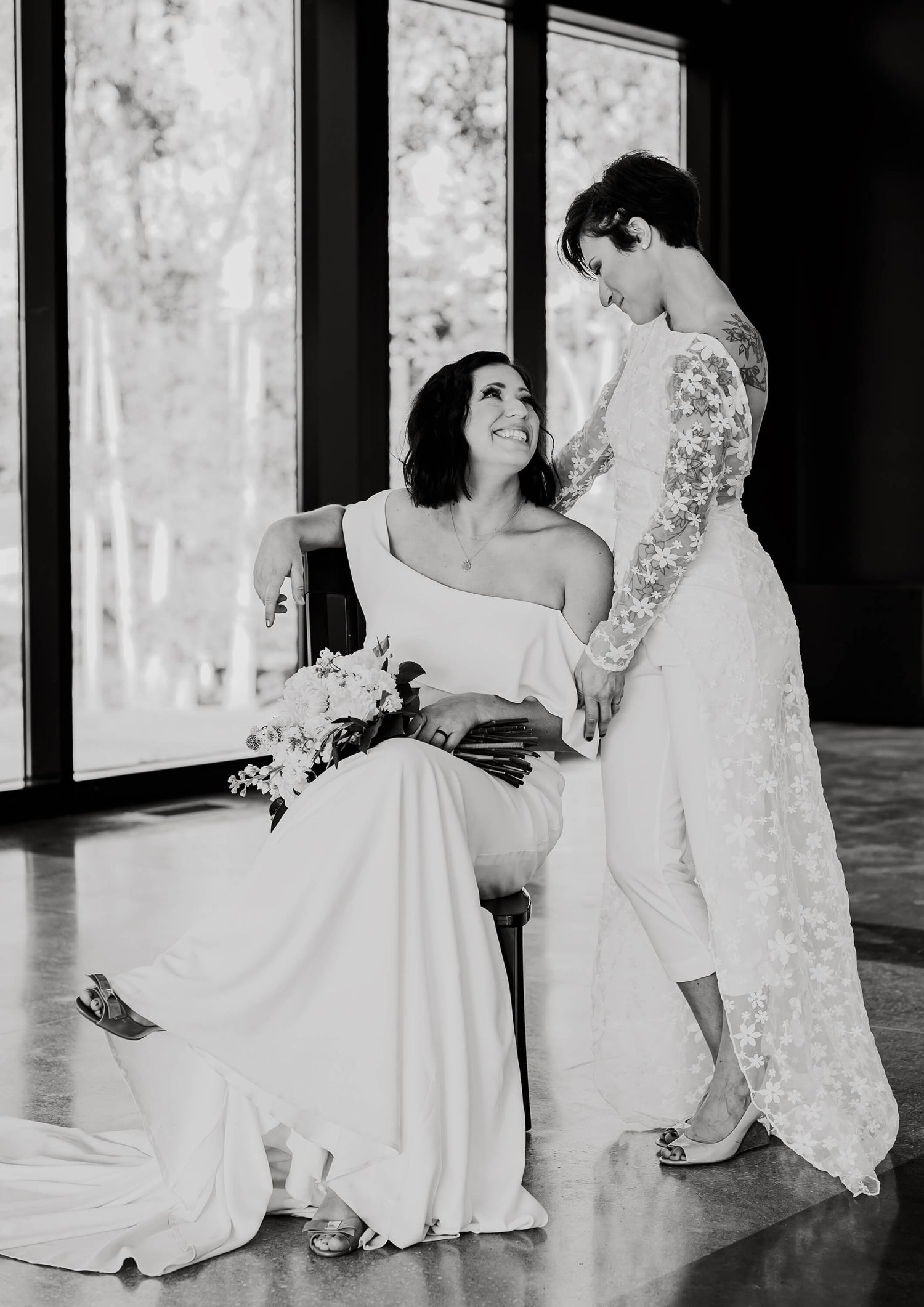 Two figures in elegant white wedding dresses share an intimate moment beside large windows in black and white photography.