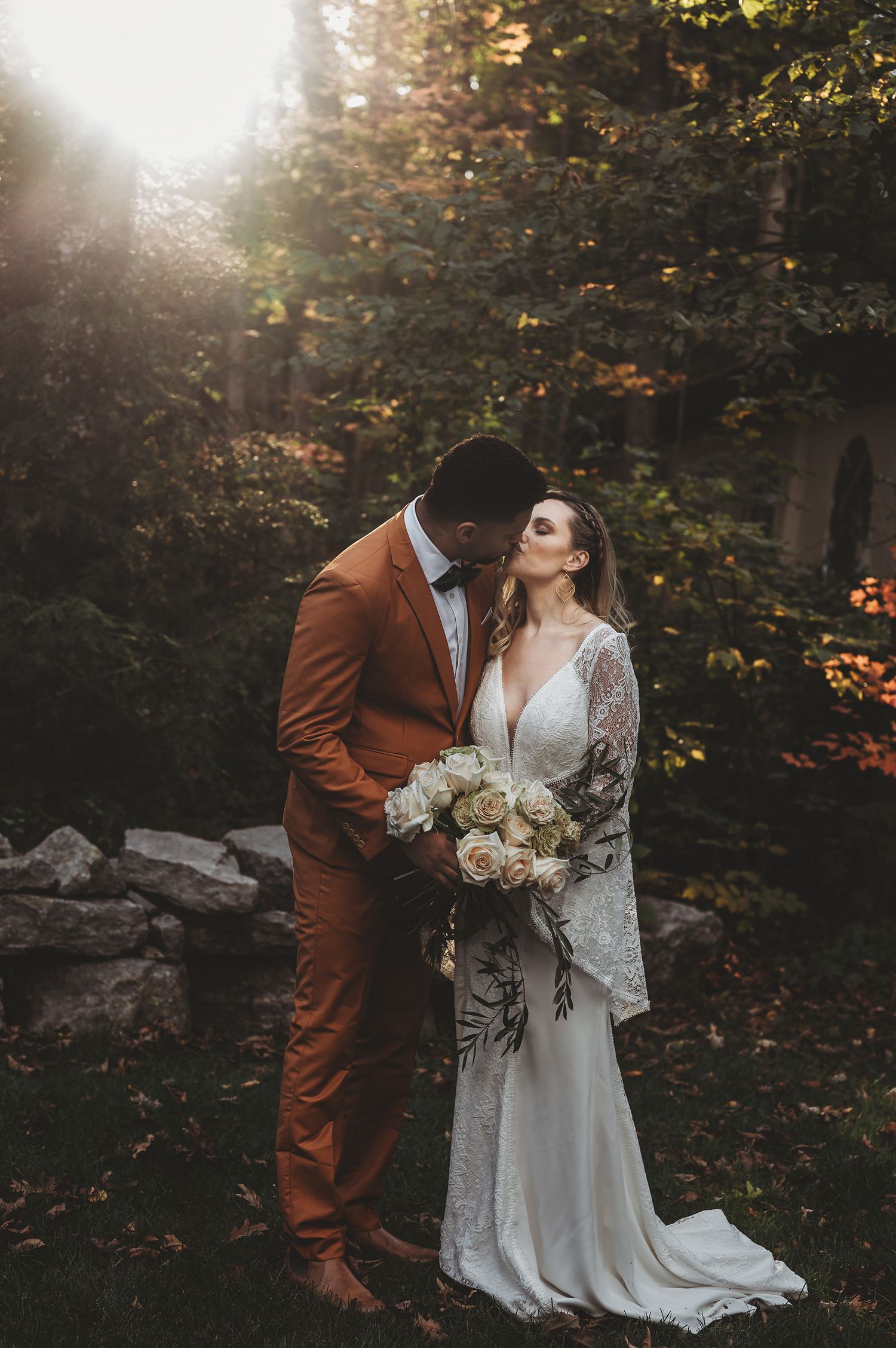 A romantic autumn wedding scene in a forest with a couple in white lace dress and brown suit sharing an intimate moment.
