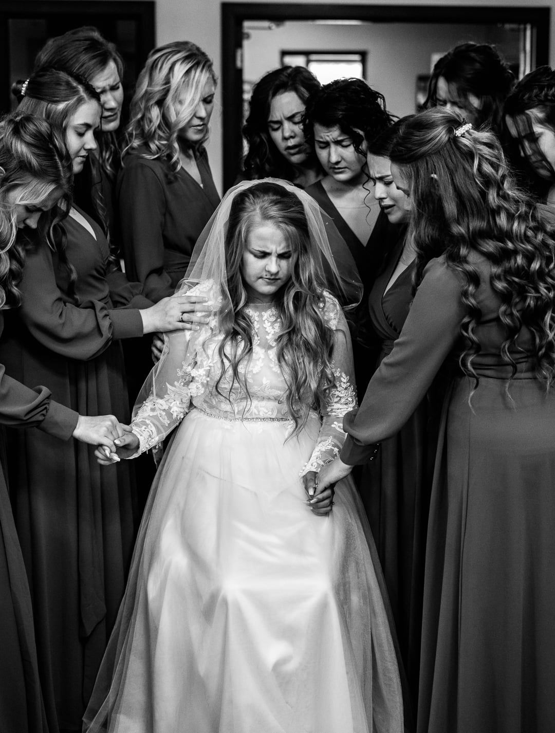 A candid black and white wedding photo showing a bride in a long-sleeved lace dress surrounded by bridesmaids in dark gowns.