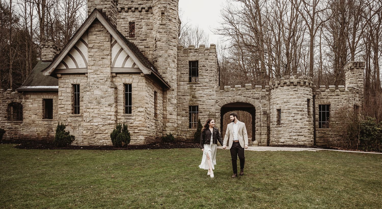 A couple walks together on the lawn in front of a gothic stone castle with towers and archways in a sepia-toned photo.