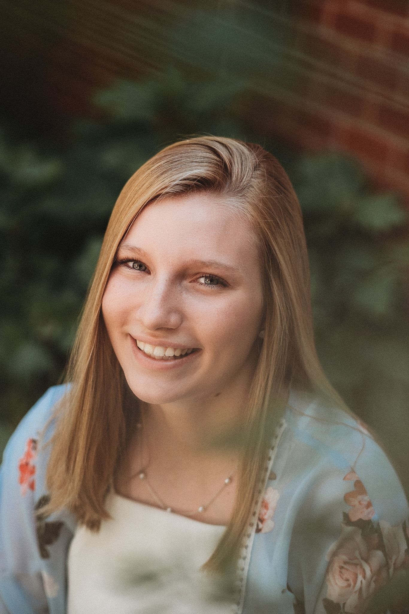 Smiling person wearing a light blue floral top poses outdoors against a blurred brick and foliage background.