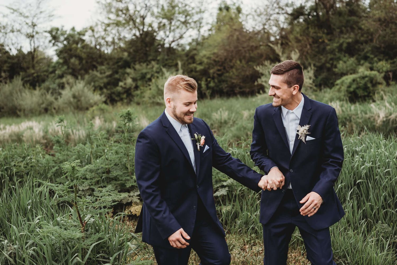 Two people in navy blue suits share a joyful candid moment while walking through tall grass in an outdoor setting.