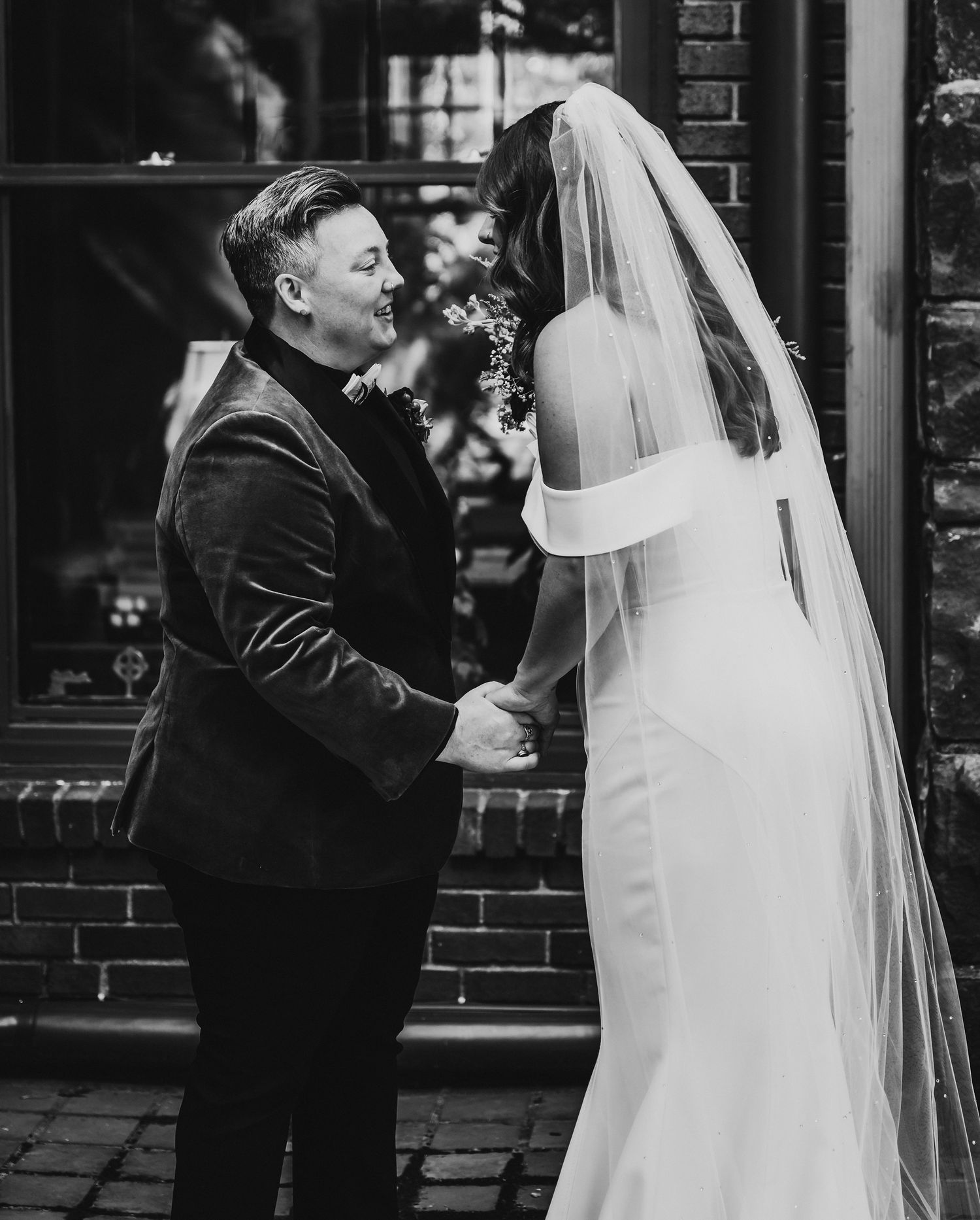 Black and white photo of couple standing together on their wedding day with elegant gown and dark suit against brick wall.