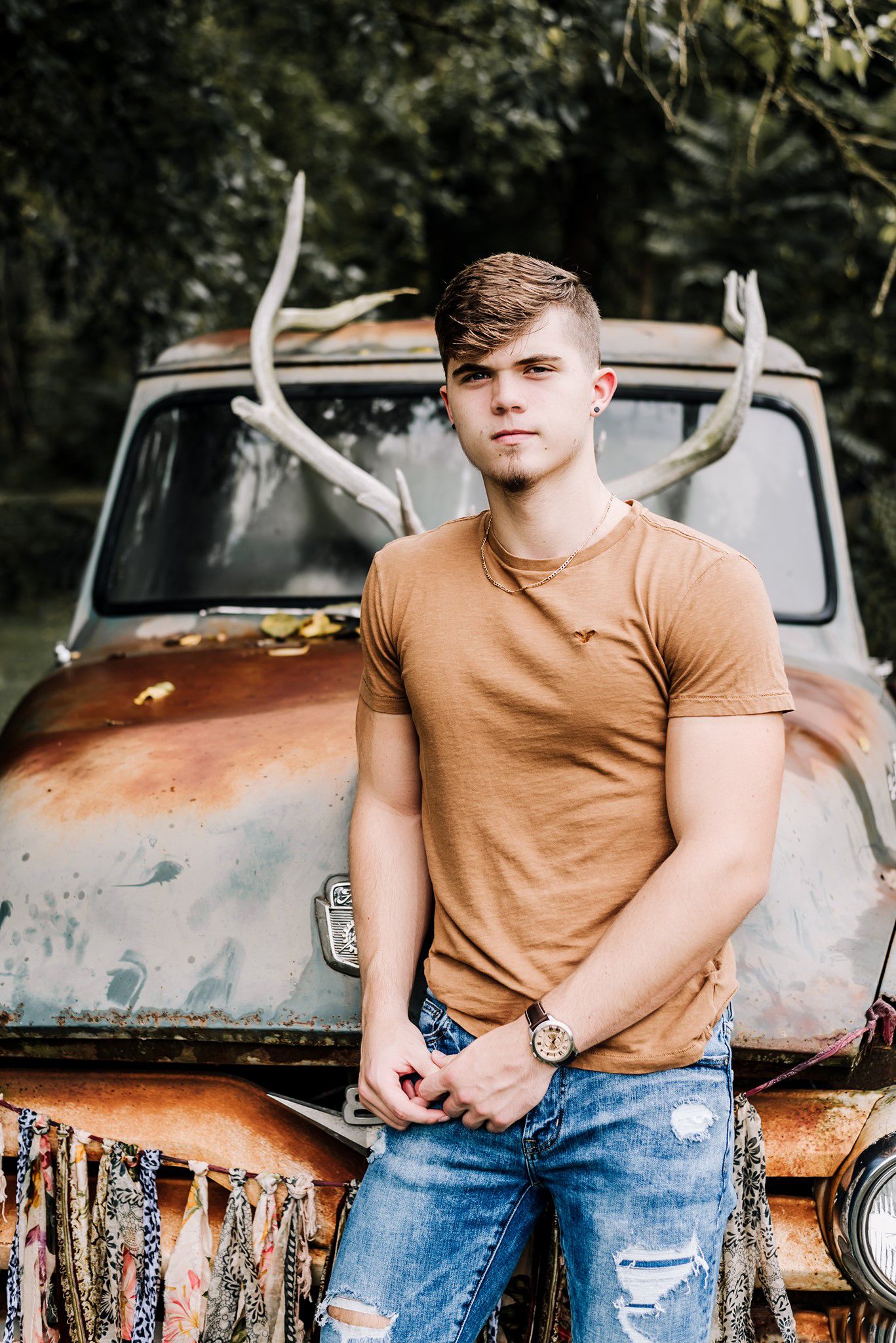 Person in tan t-shirt and distressed jeans poses against rustic vintage truck with antlers mounted on top.