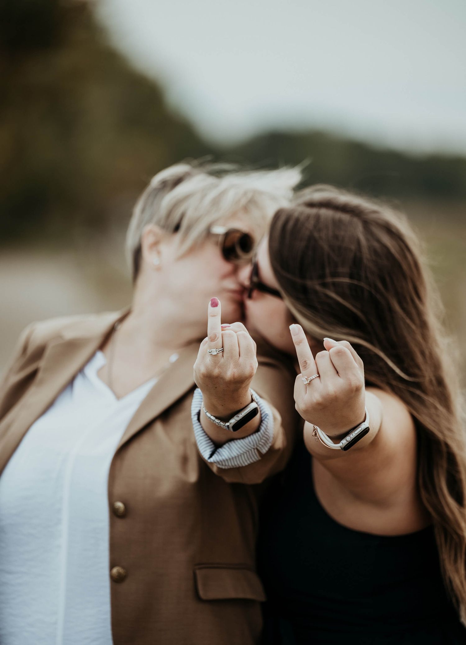 Two people in casual clothing pose together while defiantly raising their middle fingers at the camera in an outdoor setting.
