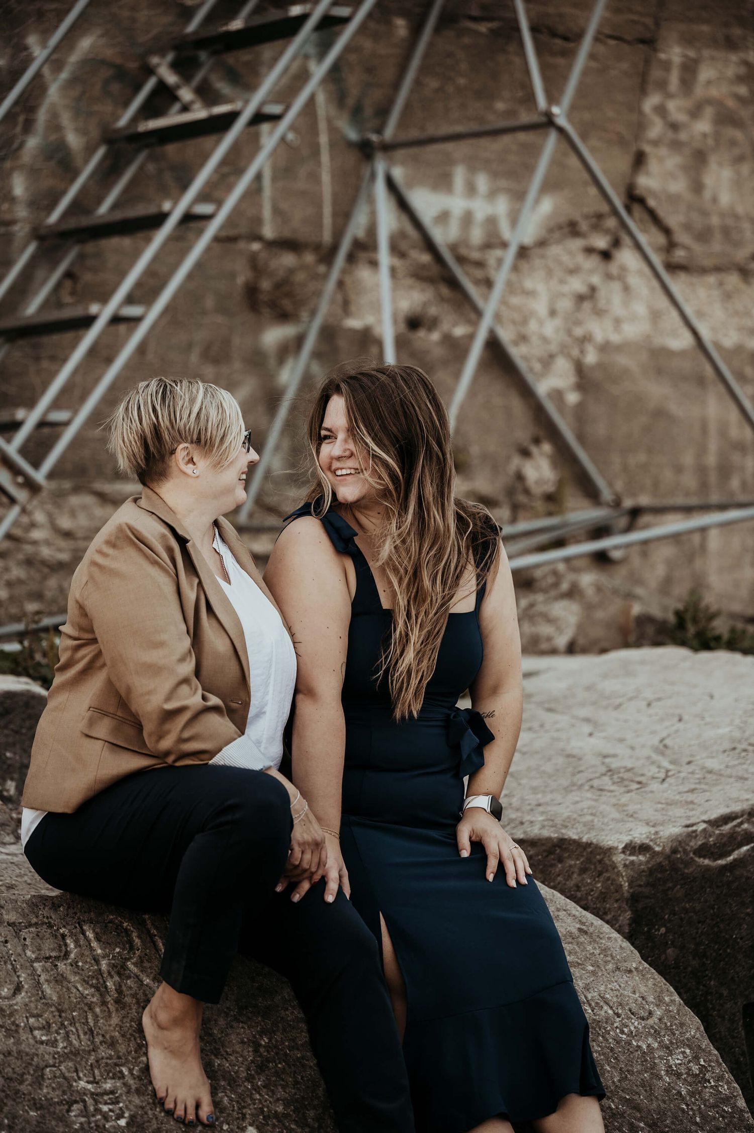 Two women sitting close together on a large rock in front of scaffolding, sharing an intimate moment at dusk.