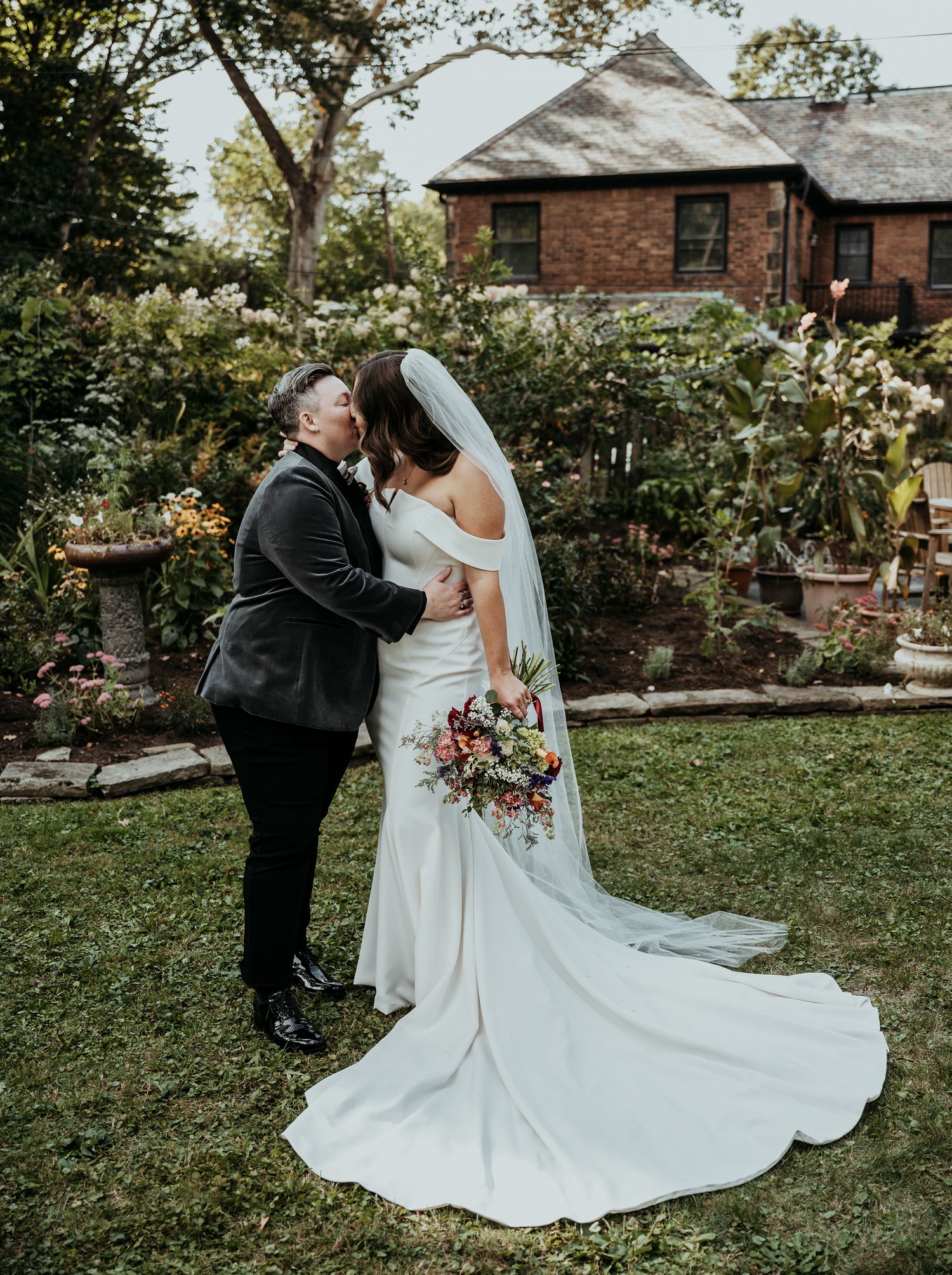Newlywed couple share an intimate moment in a garden setting with a beautiful brick house and flowing white wedding dress.