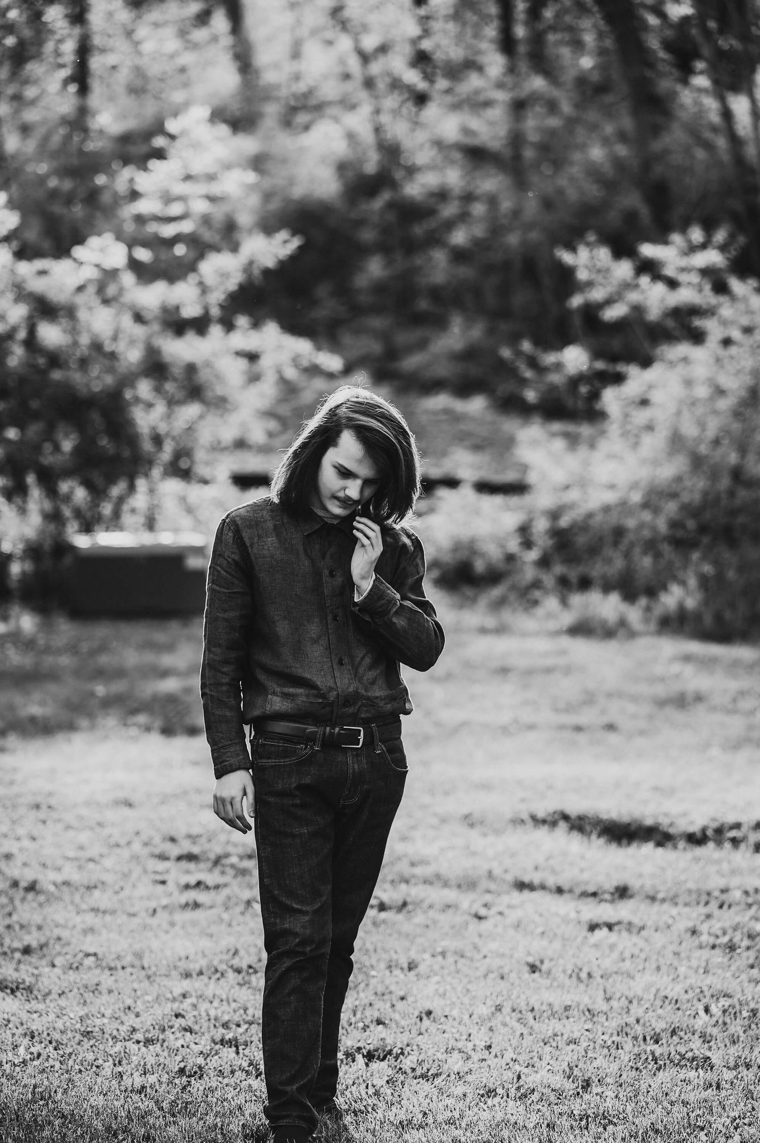 Black and white portrait of a person in a hoodie and jeans standing outdoors with trees in background.
