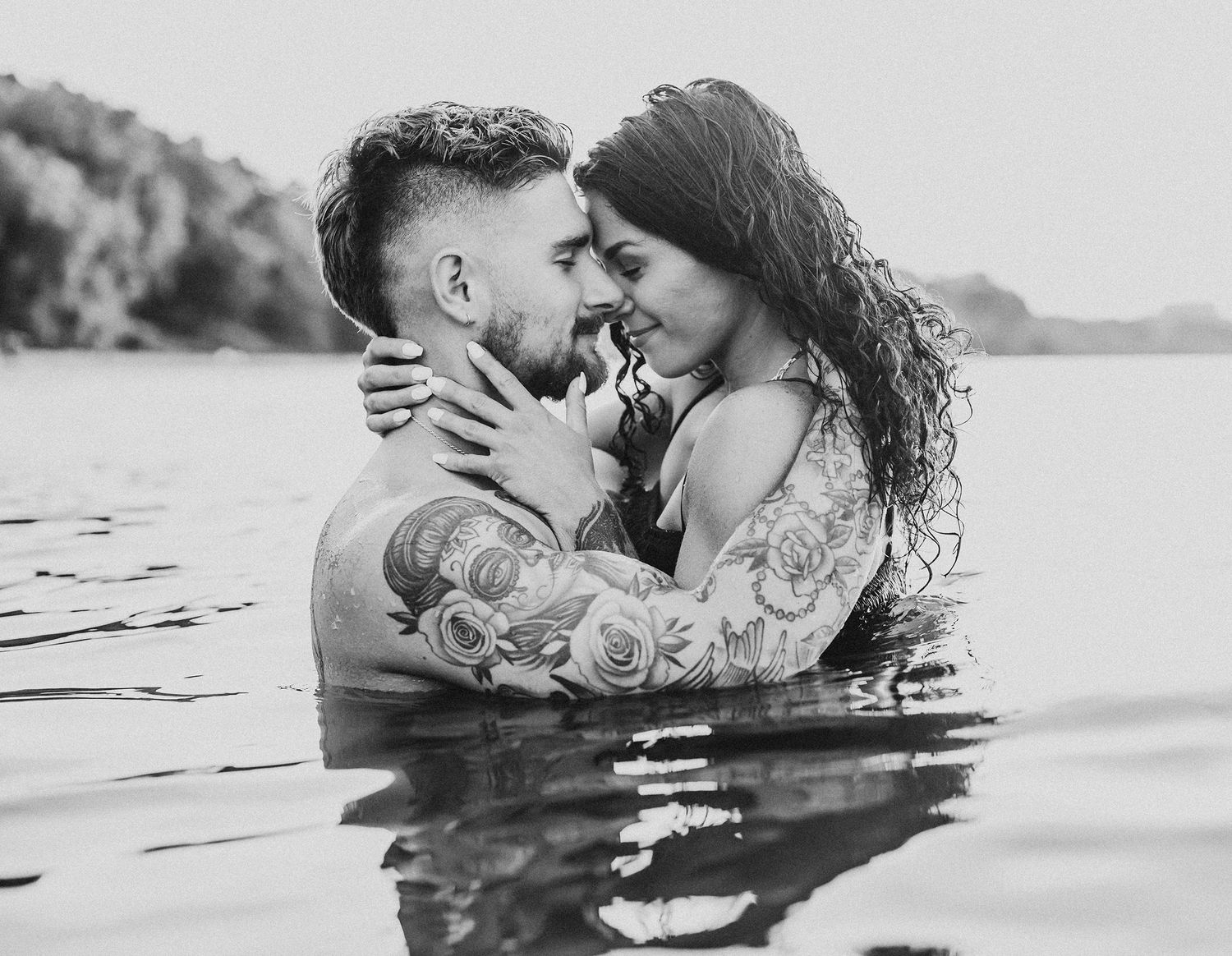 A romantic couple embraces while standing in water, captured in black and white photography at a lake.