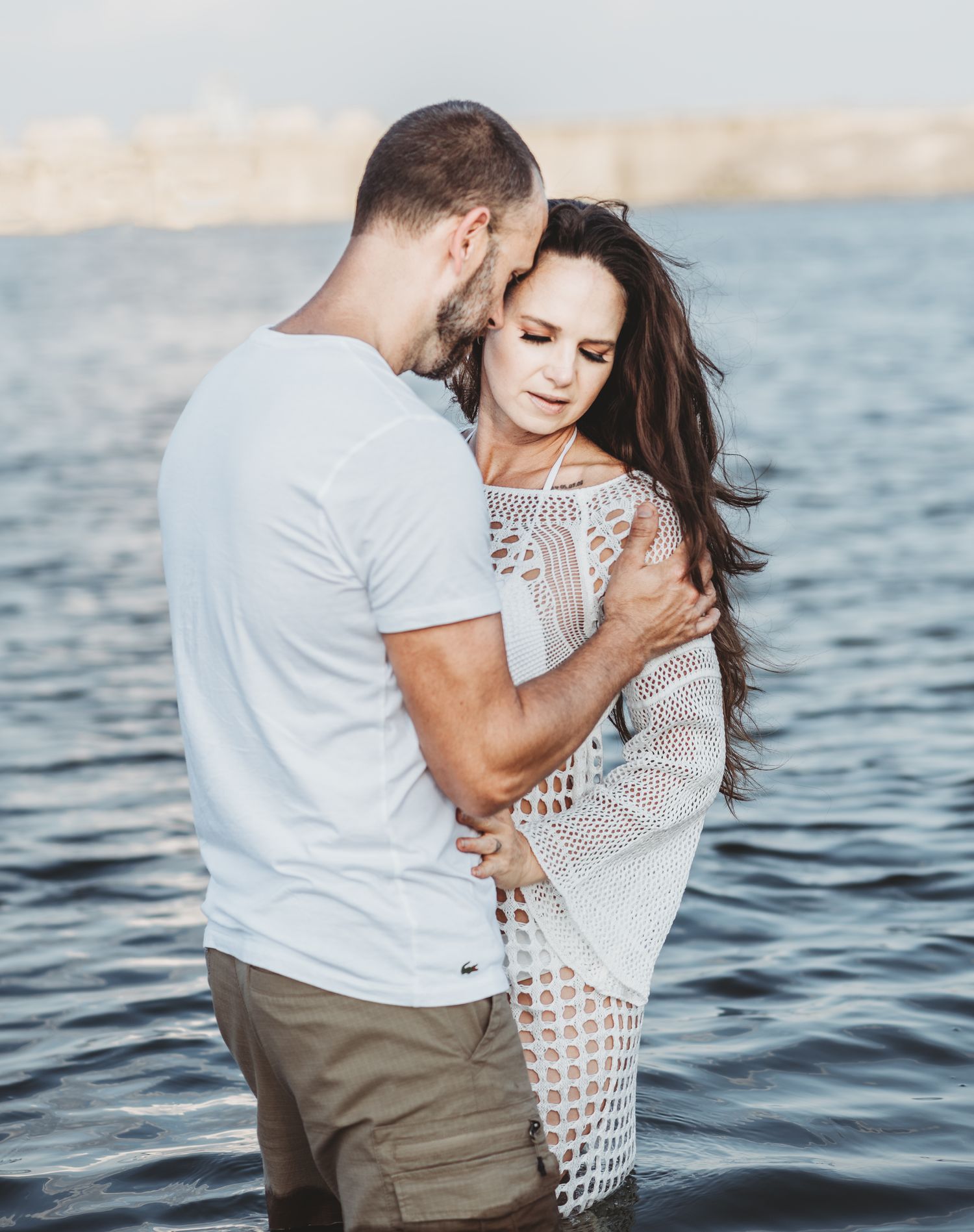 A romantic couple embraces by the ocean at sunset, with the water glistening behind them.