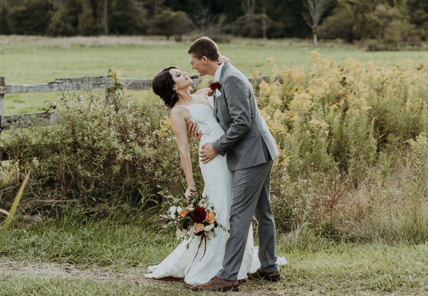 A couple shares a romantic dance pose in a grey suit and white dress with a bouquet among wildflowers in a field.