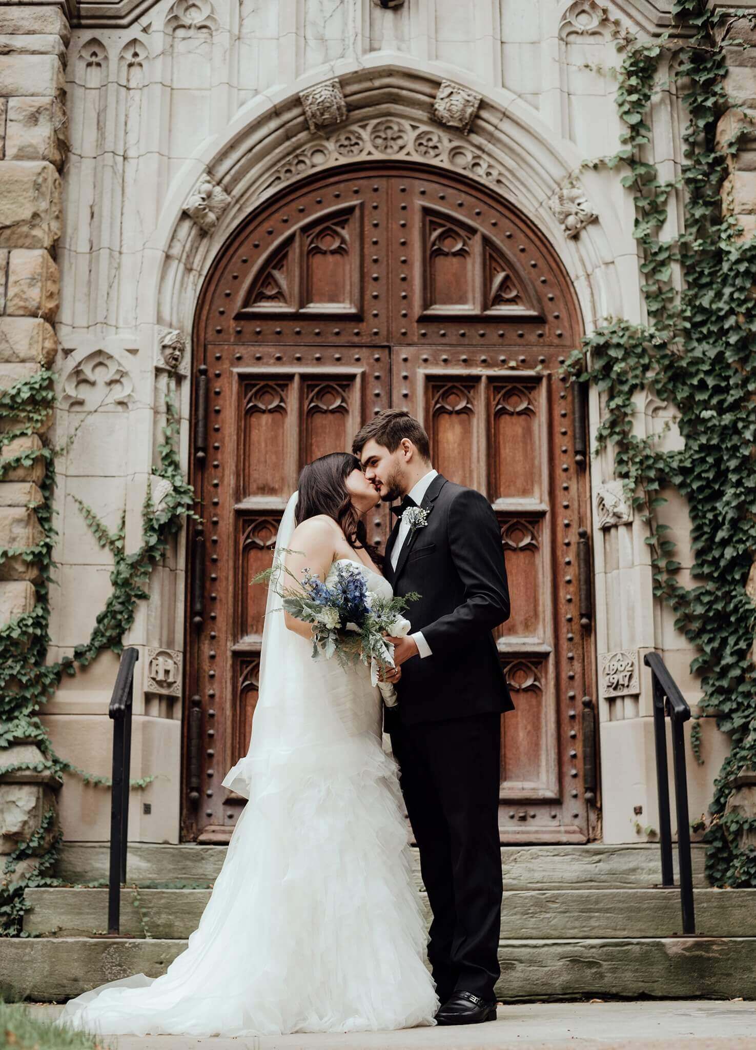 Couple sharing intimate moment in front of ornate wooden church doors with ivy climbing stone archway.