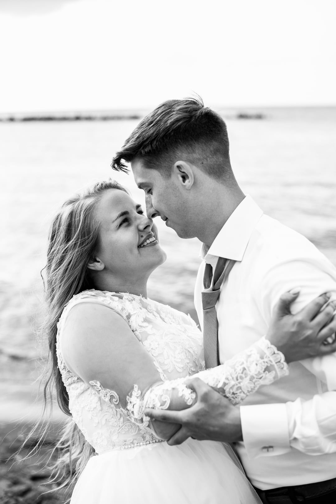 Black and white romantic photo of newlyweds dancing and embracing on a beach, with the bride in a lace wedding dress.
