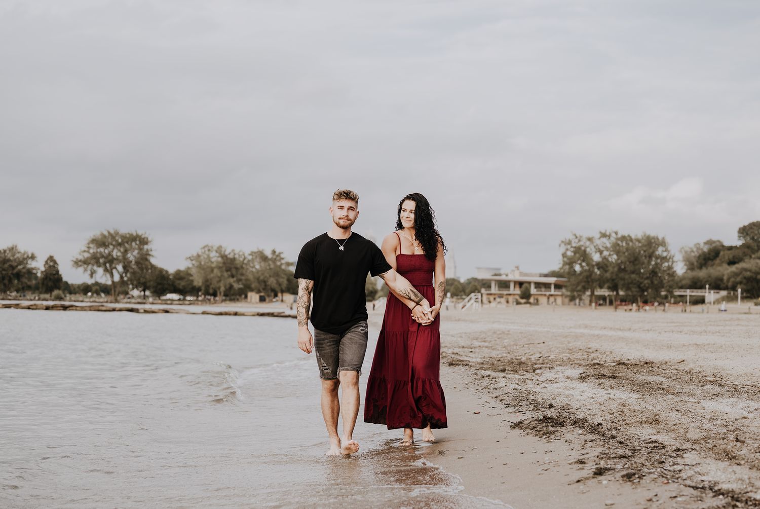 Couple walking barefoot together along a scenic beach shoreline on an overcast day.