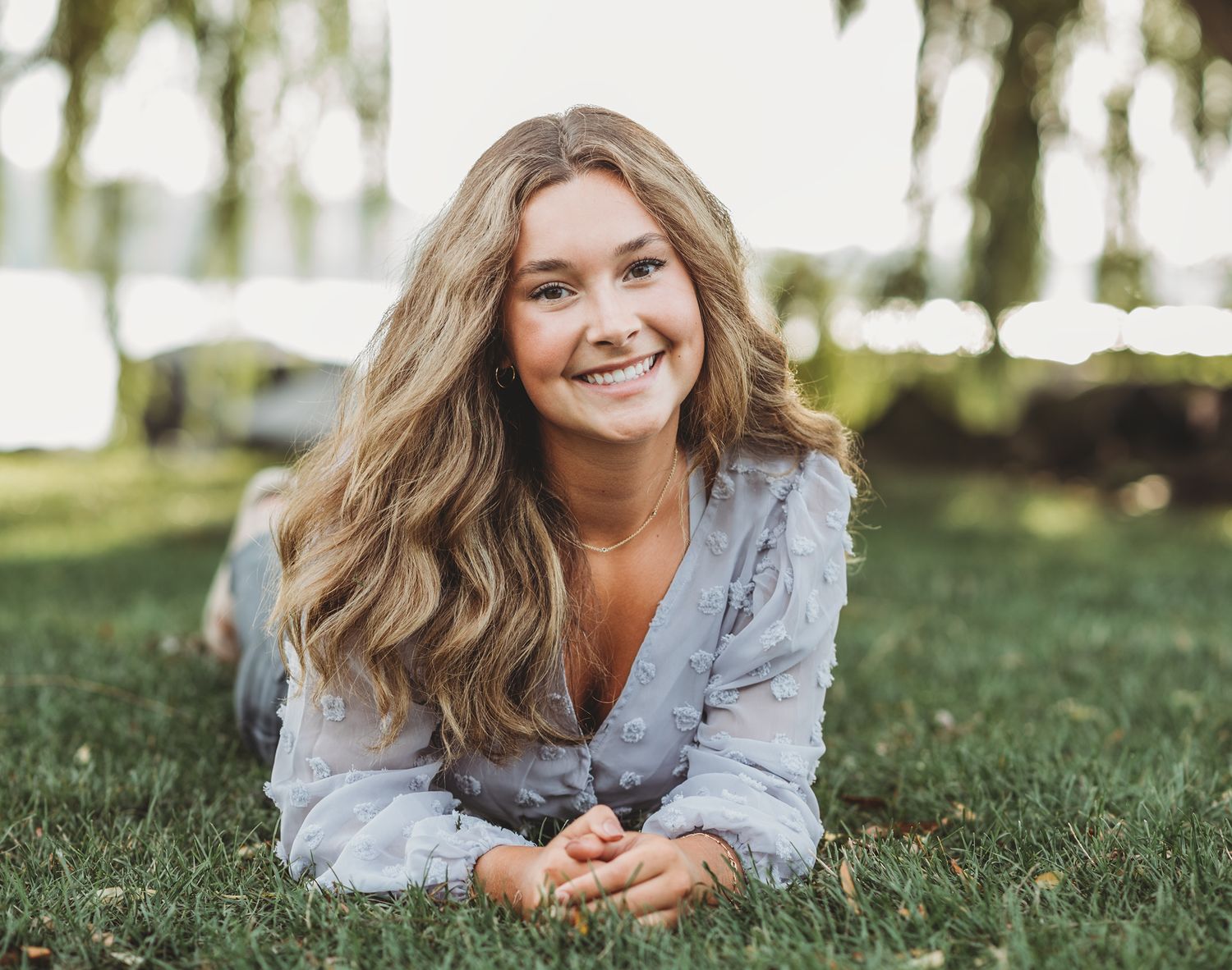 Someone with flowing wavy hair lies on grass smiling at the camera while wearing a light denim shirt on a sunny day.