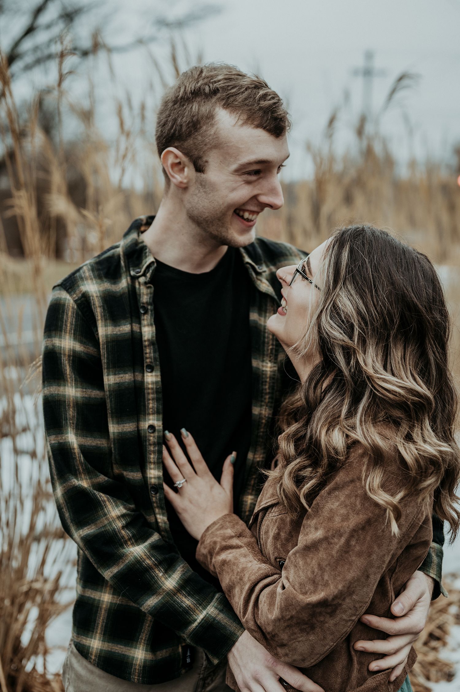A romantic couple laughs and embraces while wearing casual winter clothing in a field during golden hour.