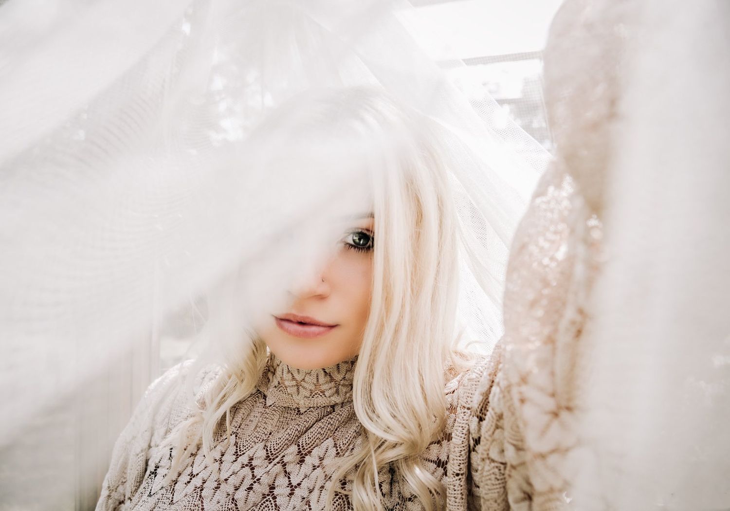 A person with platinum blonde hair looks through sheer white fabric while wearing a textured sweater.