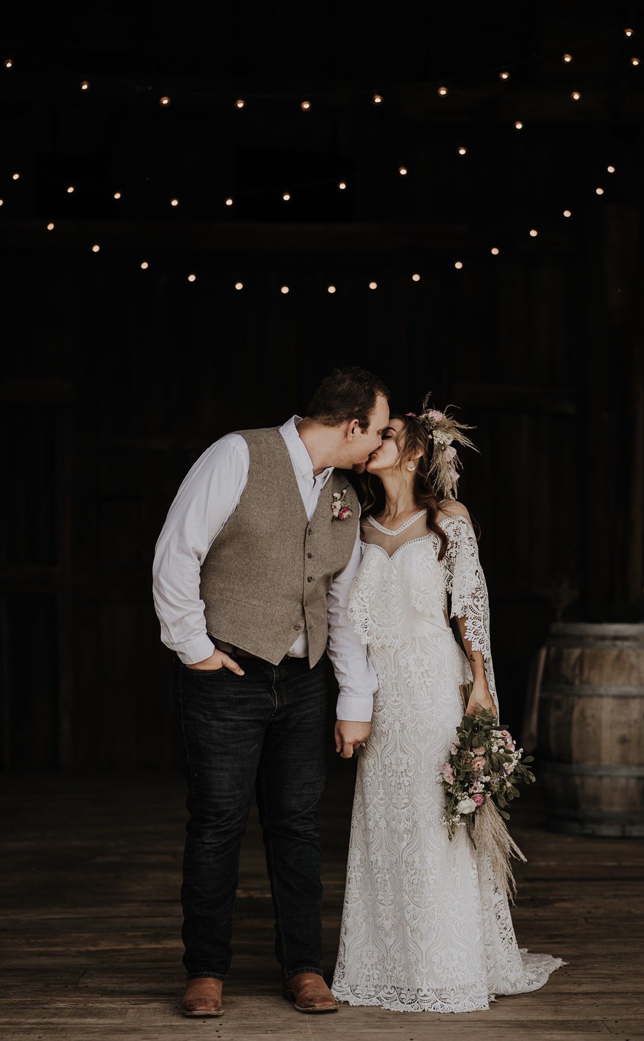 Newlyweds share an intimate moment under string lights on a rustic wooden deck with wine barrels in the background.