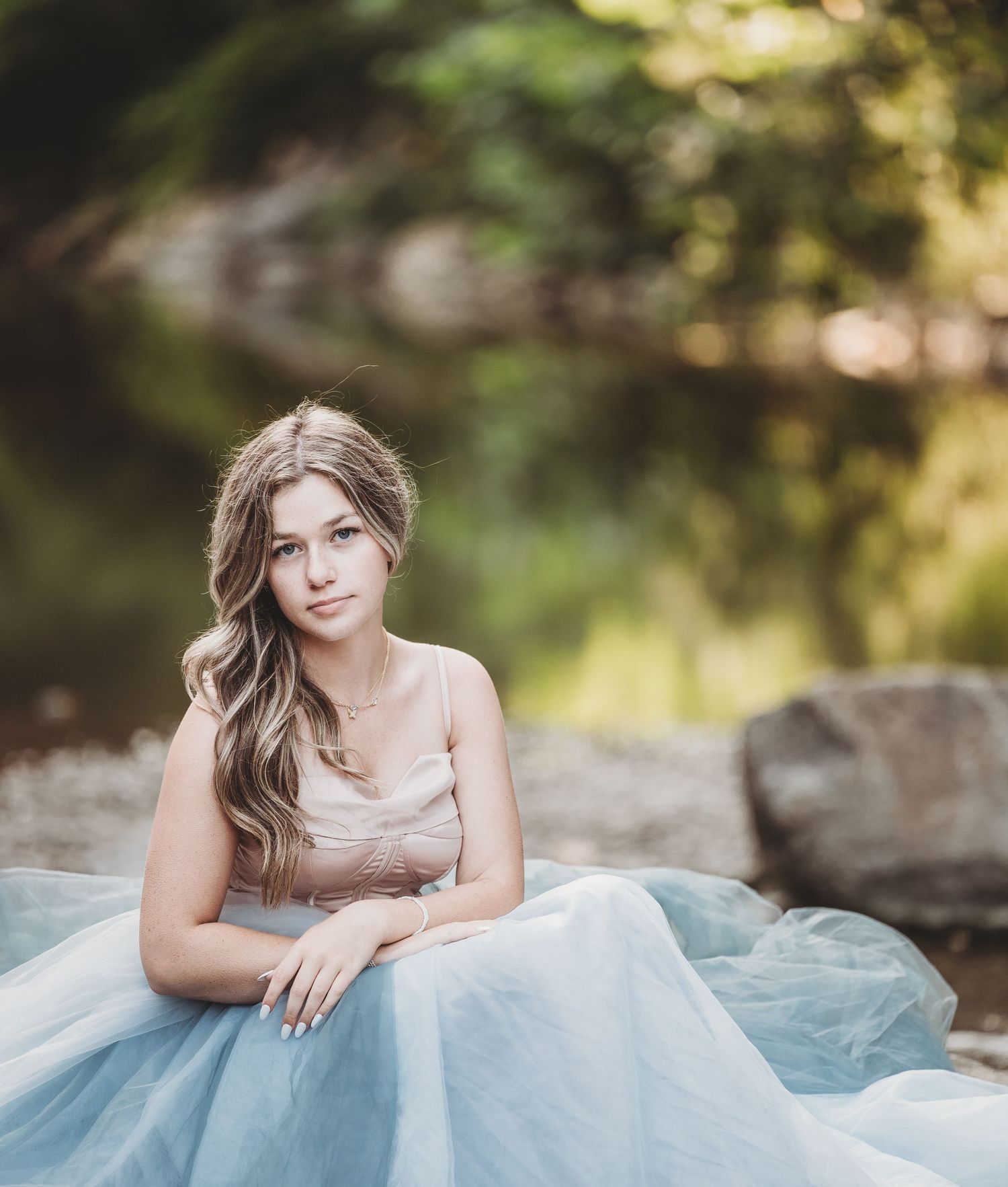 Young woman in light blue tulle gown poses gracefully against natural outdoor backdrop.
