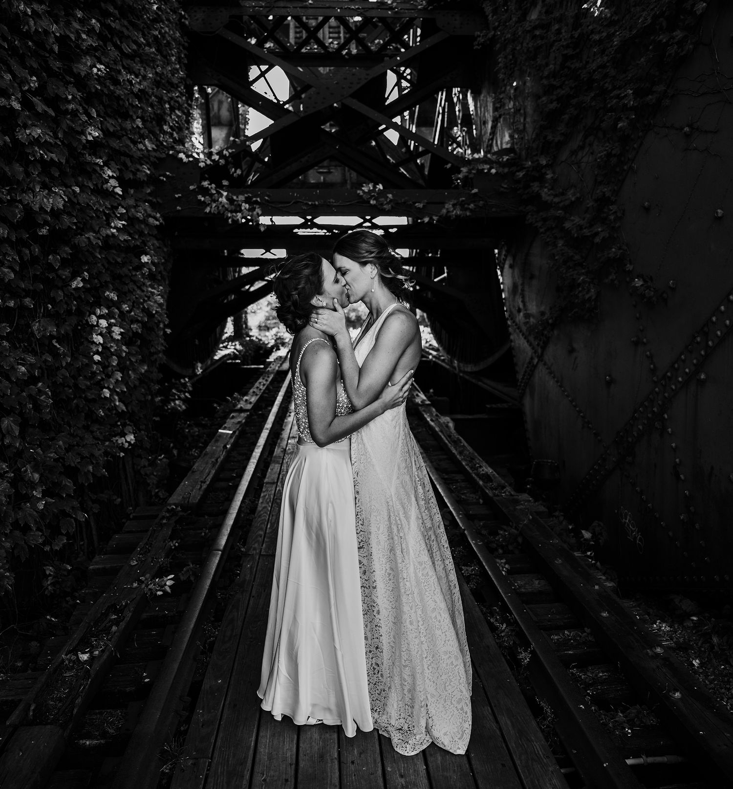 A couple embraces in a romantic black and white moment on a moody wooden bridge covered in vines.