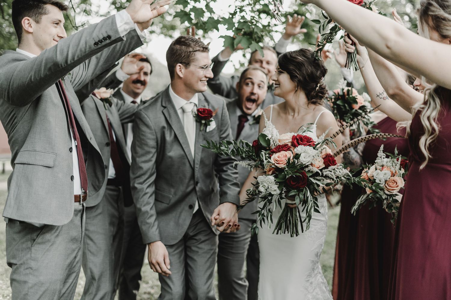 Wedding party members in grey suits and burgundy dresses celebrate with champagne outdoors.