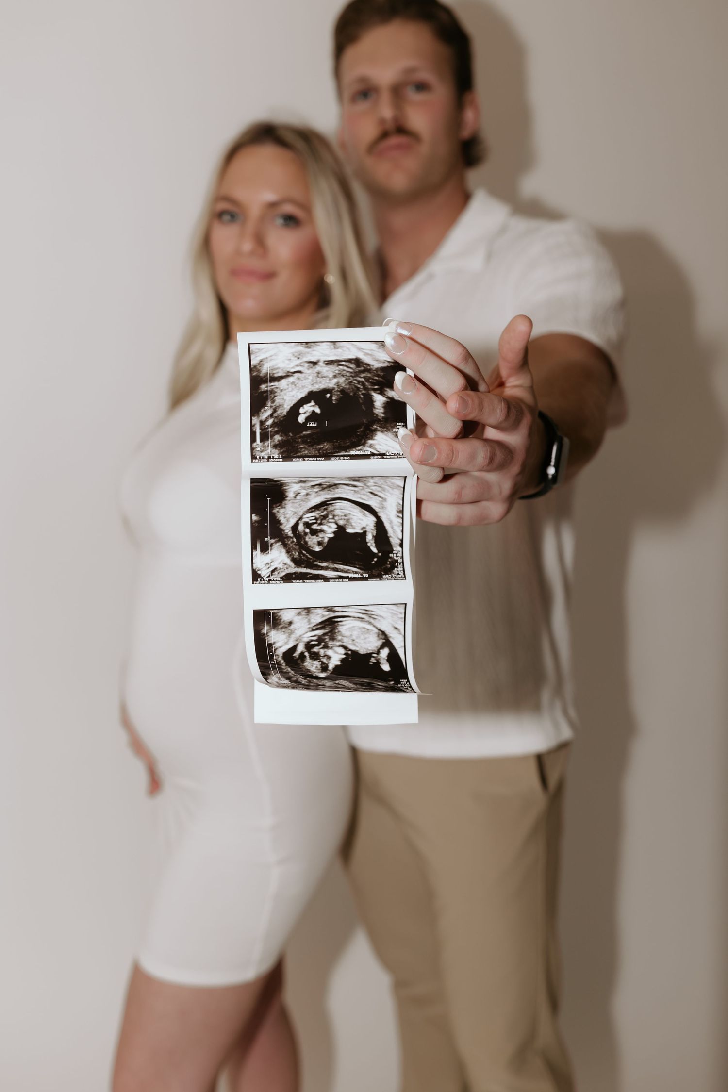 A couple in white and beige clothing displaying ultrasound images against a white wall.