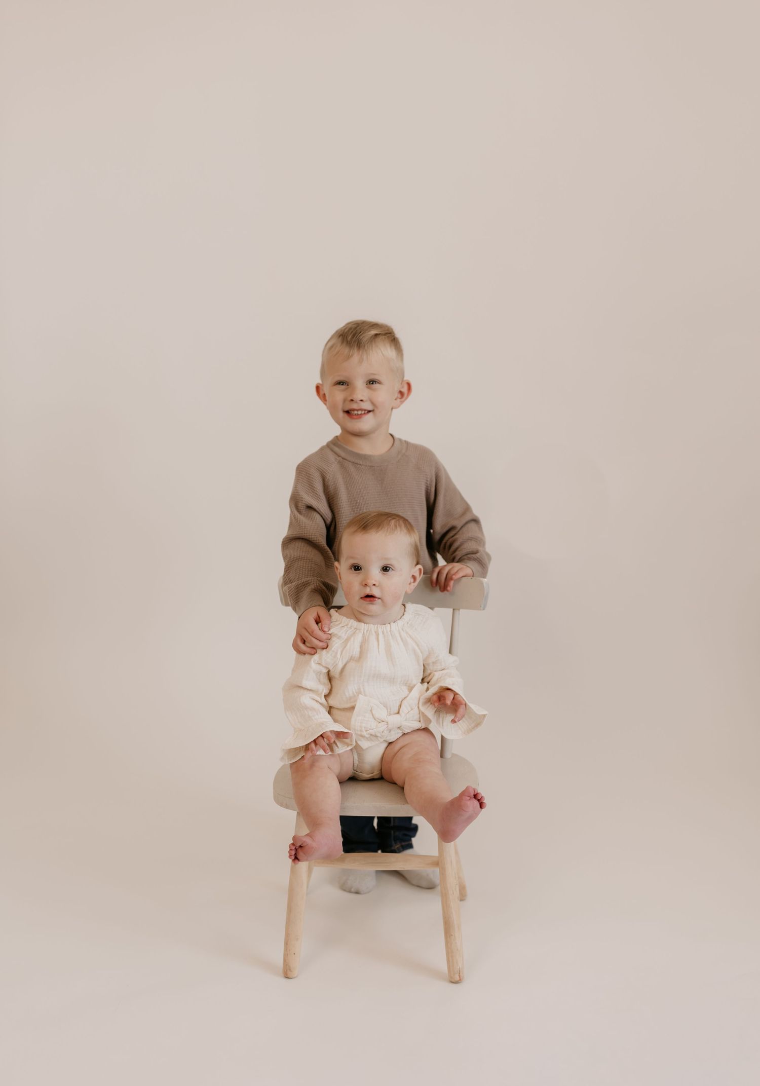 Two children posing together on wooden stool against neutral backdrop.