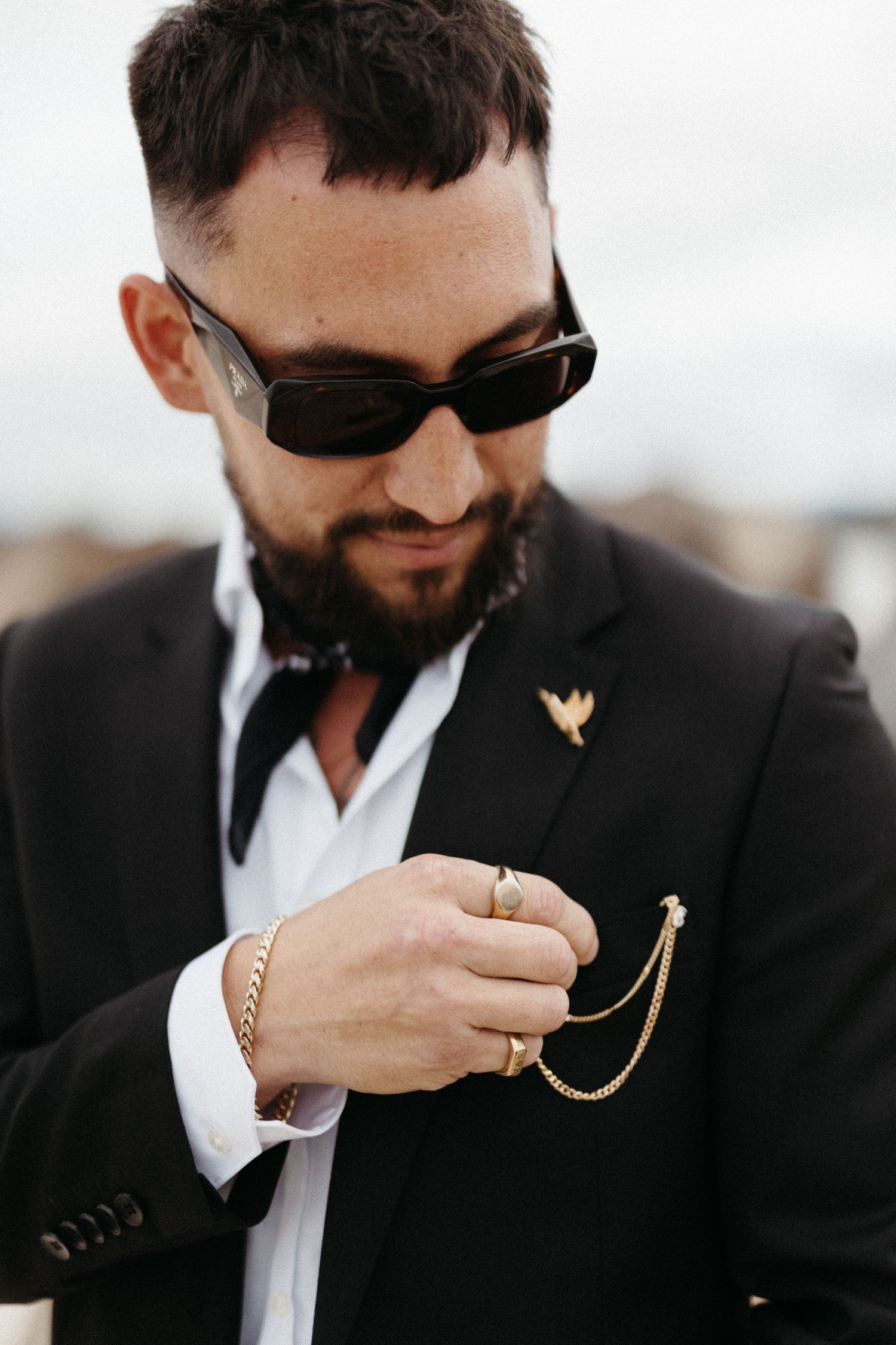 Stylish groom in black suit and sunglasses adjusts gold chain accessory on lapel against light background.