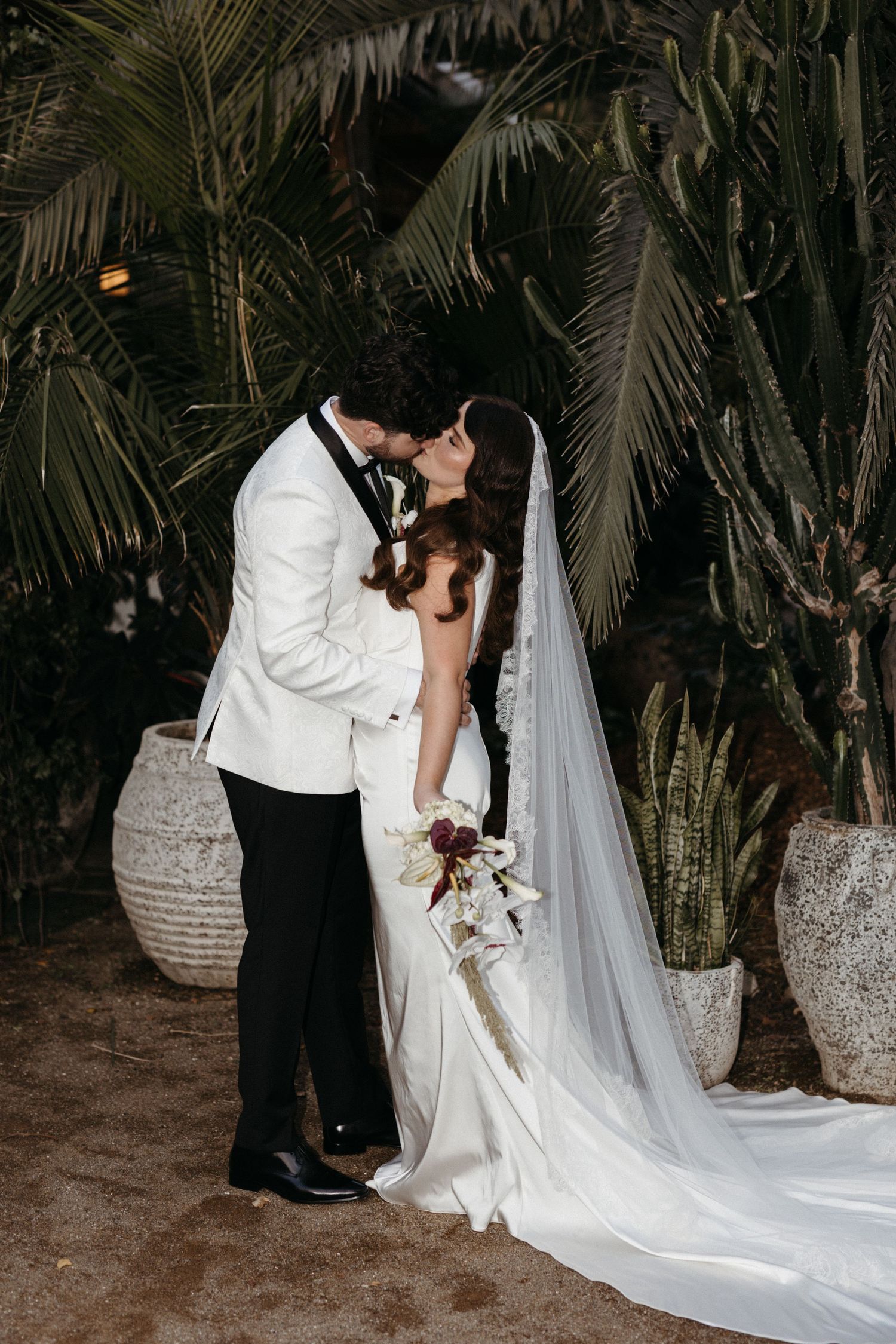 A romantic wedding scene outdoors at night showing a couple in formal attire with a flowing white veil and palm trees behind.