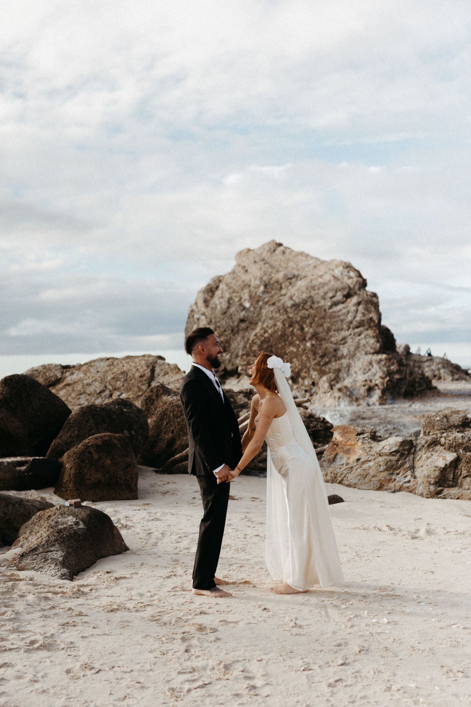 A couple walks along a sandy beach with large rocky outcrops and moody cloudy skies in a portrait series.