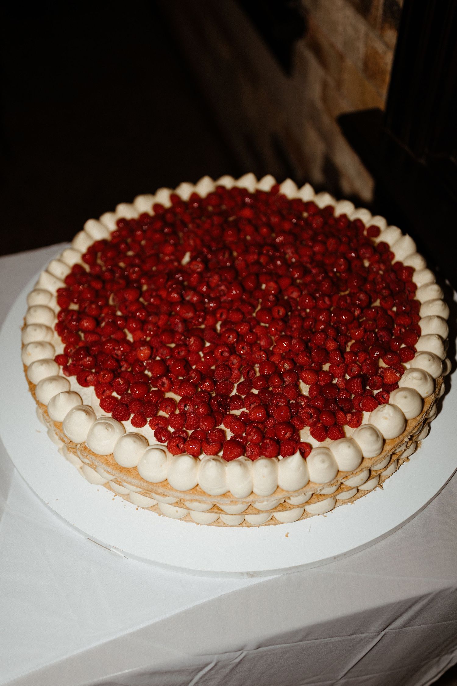 A beautiful cake decorated with fresh raspberries and piped white cream border sits on a white tablecloth.